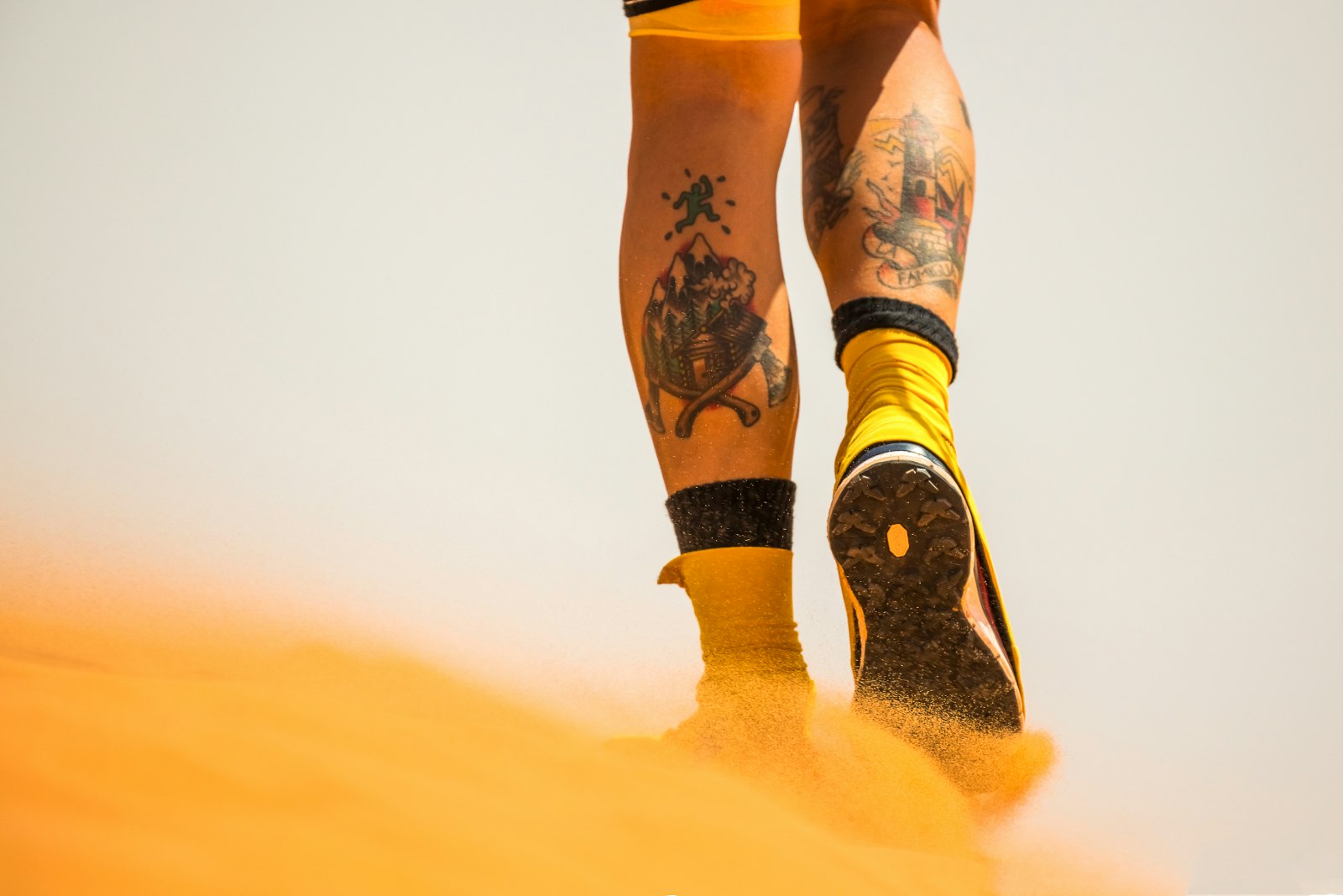 The image features the lower legs of an ultra runner from behind; the calf muscles both have large tattoos on them. Wind blows sand to the left that has been kicked up by the runner's foot
