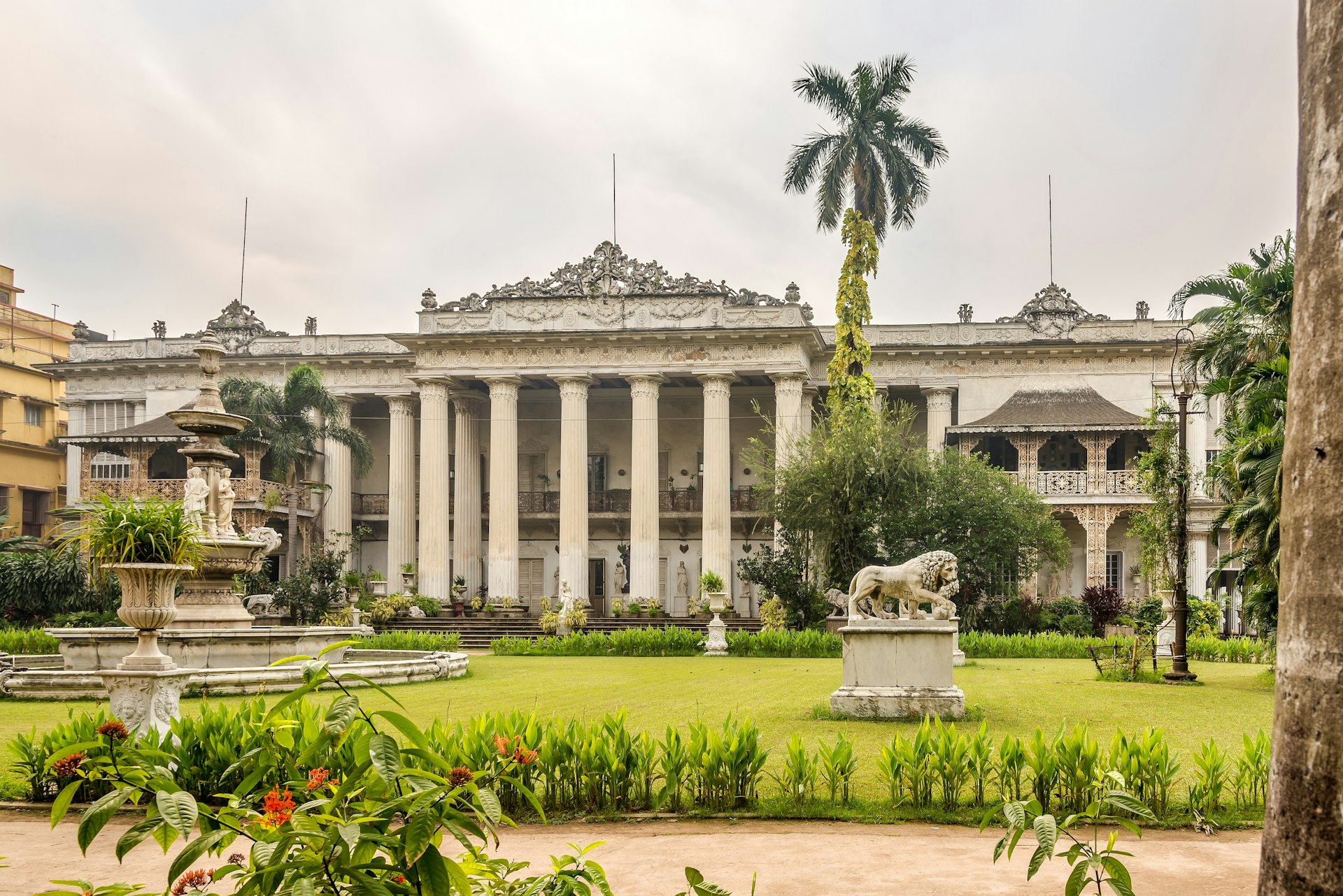 The exterior of Kolkata's Marble Palace as seen from the front. The palace is grand, with white columns and fountains in the green gardens.