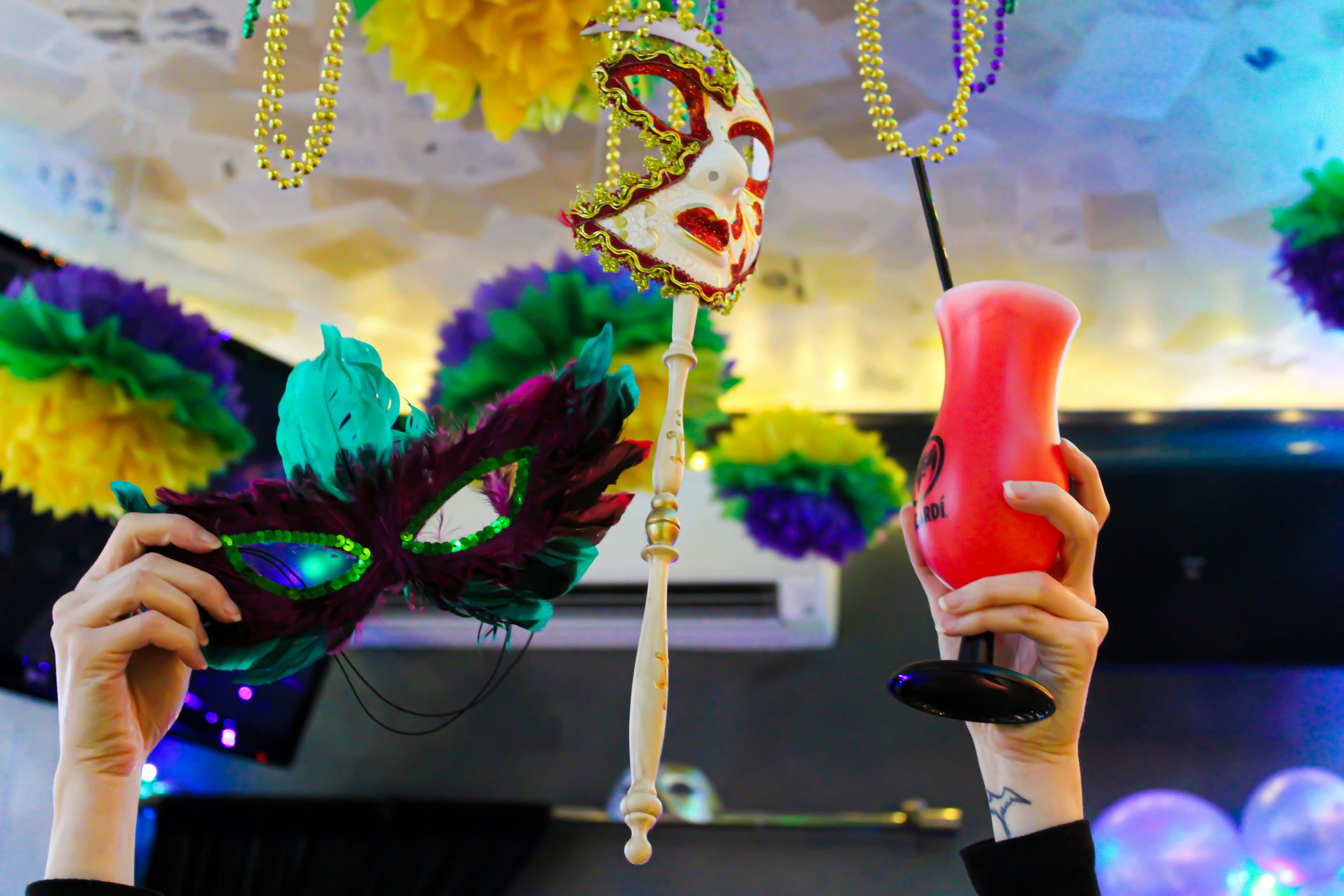 Hands hold a cocktail and mask in the air, with beads in the background