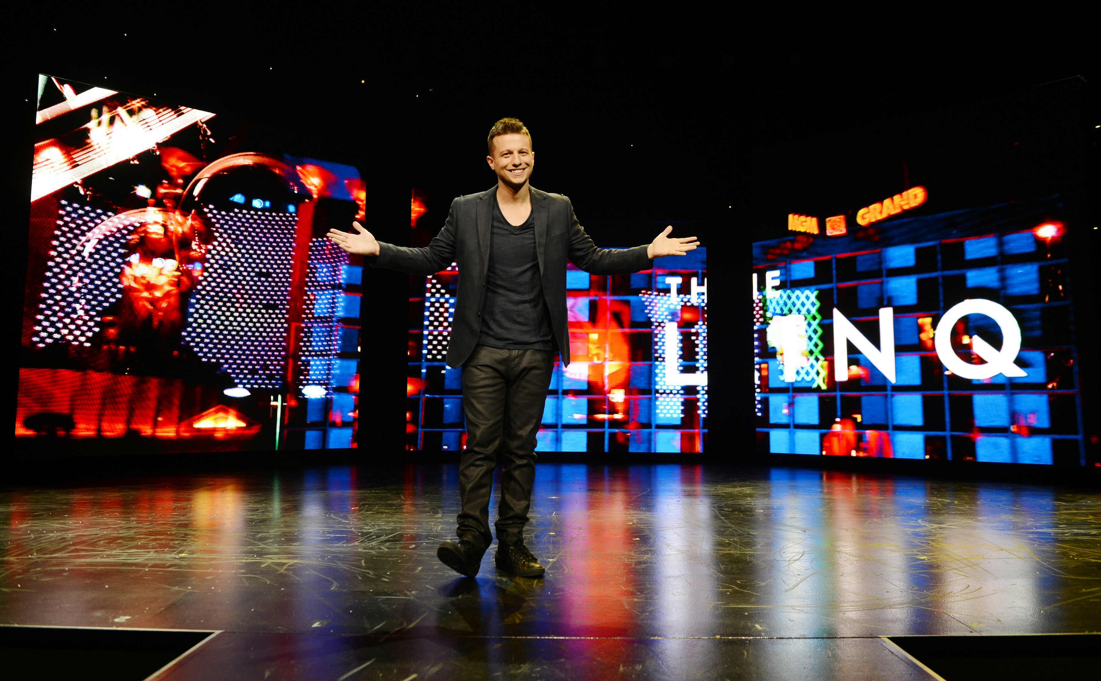 Magician Mat Franco, dressed all in black, stands on stage in Las Vegas, smiling and holding out his arms in a welcoming pose.