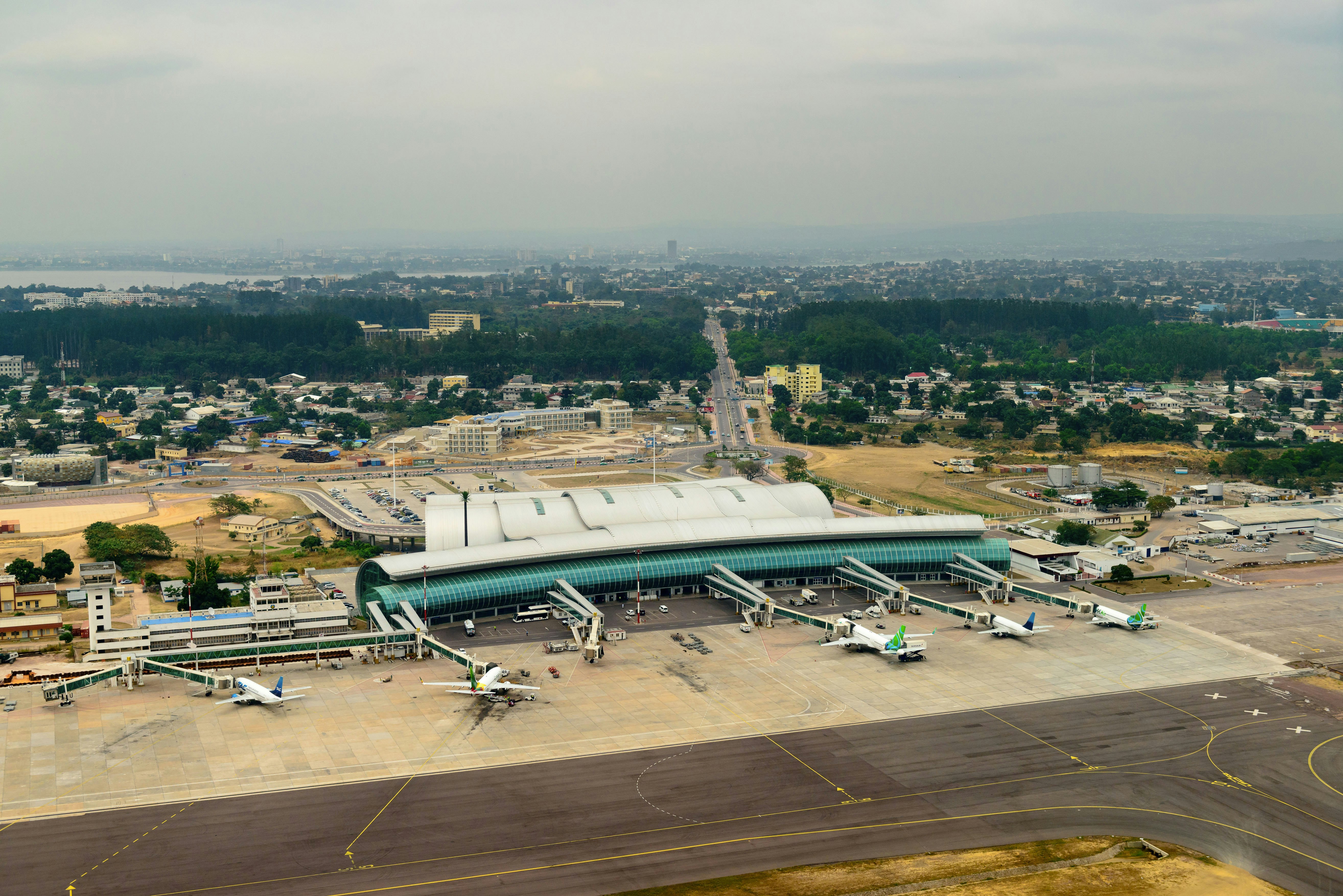 An exterior shot of the Maya Maya Airport in Brazzaville in the Republic of Congo shoes five planes parked outside a long silver modernist airport with a long teal awning in front. In the background is a rich forested landscape with a city beyond it and water on the horizon