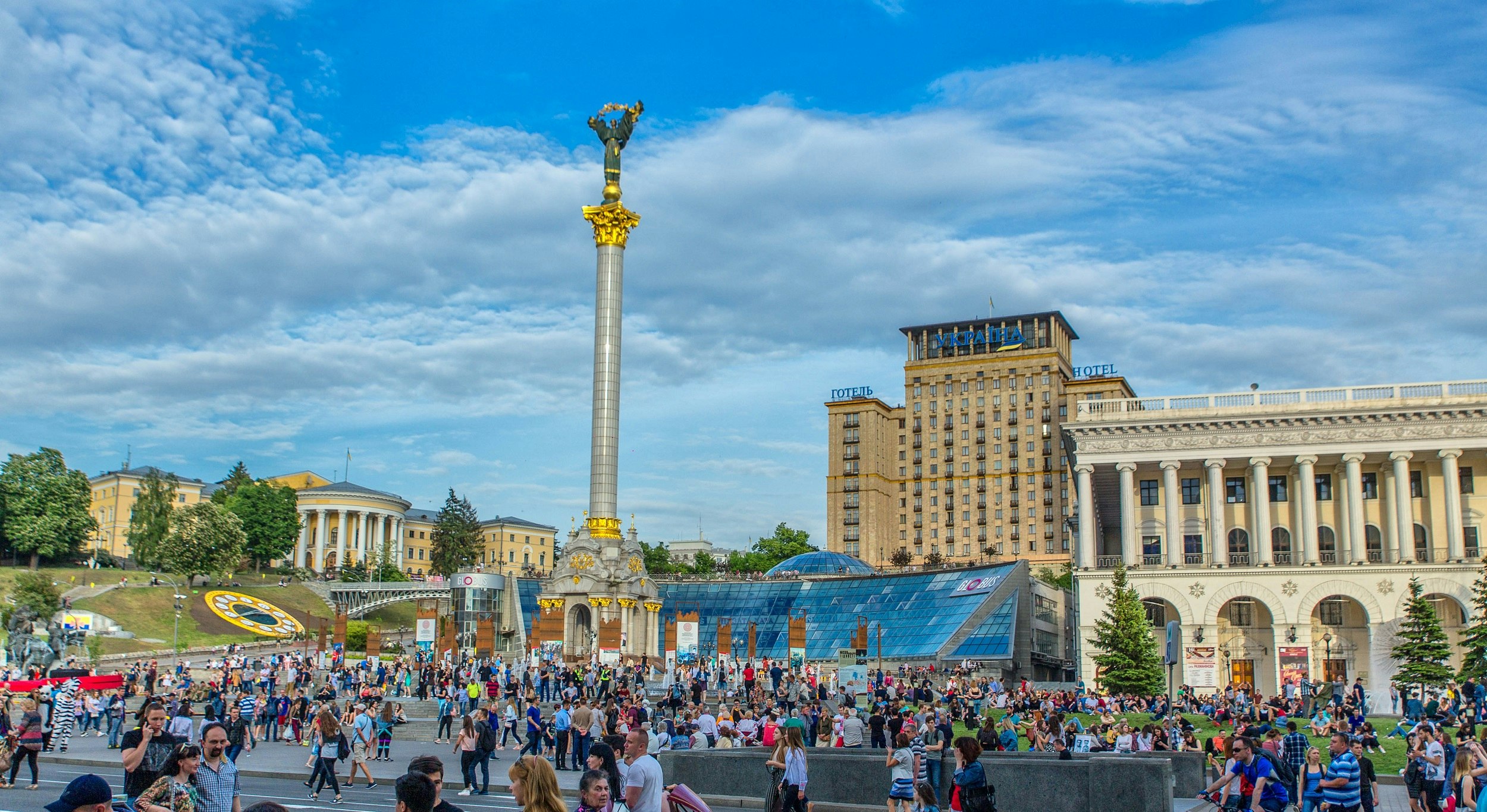 A square in the centre of Kiev is crowded with people. A large columned monument with an angel on top dominates the square