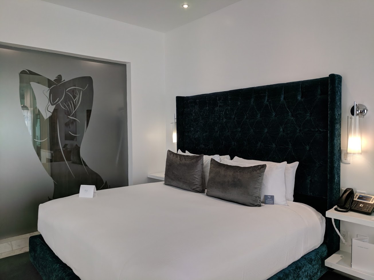 A white hotel bed in Cabo San Lucas sits by a glass partition with the stylized outline of a woman etched onto it in varying tones of grey