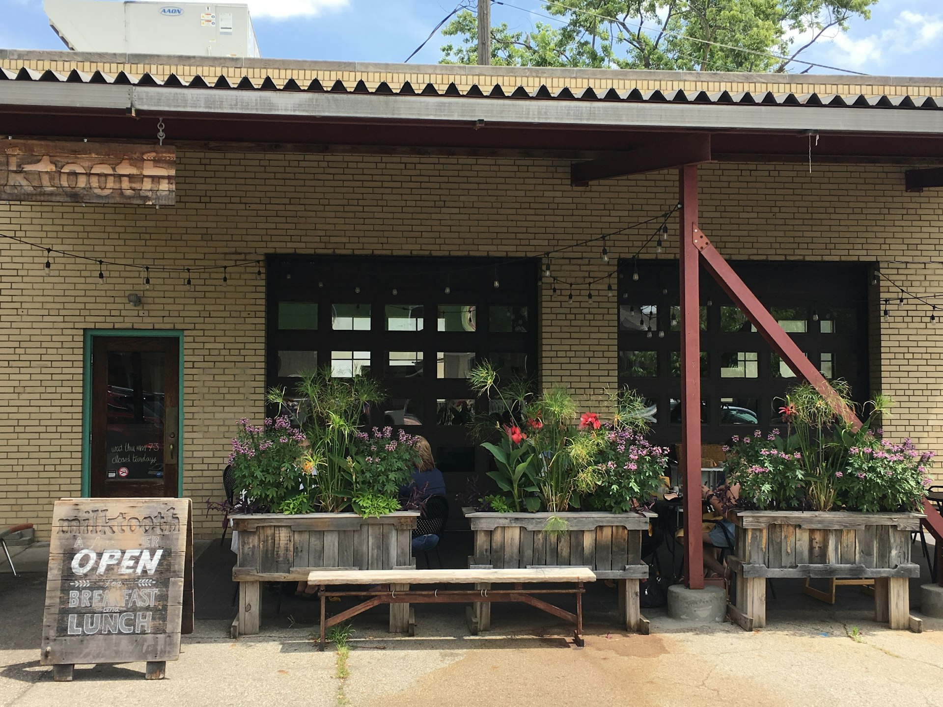 Exterior view of Milktooth with light colored bricks and wooden structures holding assorted flowers.