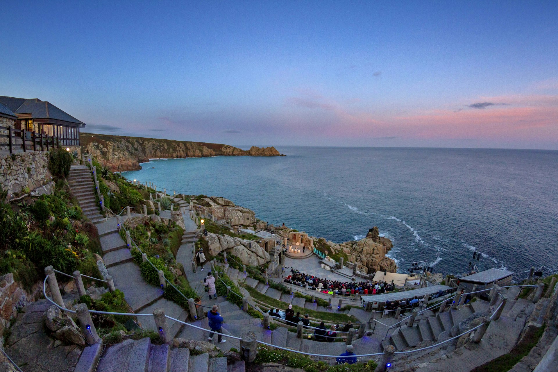 Steps lead down to the open-air Minack Theatre in Penzance; it's sunset, and the Atlantic Ocean can be seen beyond the cliff-top performance.