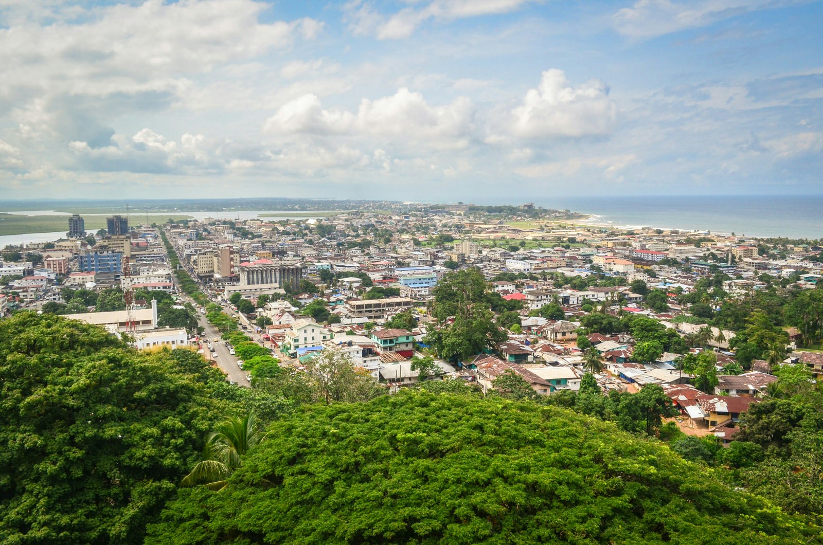 A view over the capital city of Monrovia and out to the ocean beyond; the main streets are lined with trees and the rooftops a mix of terracotta, rusted corrugated metal and silver tin