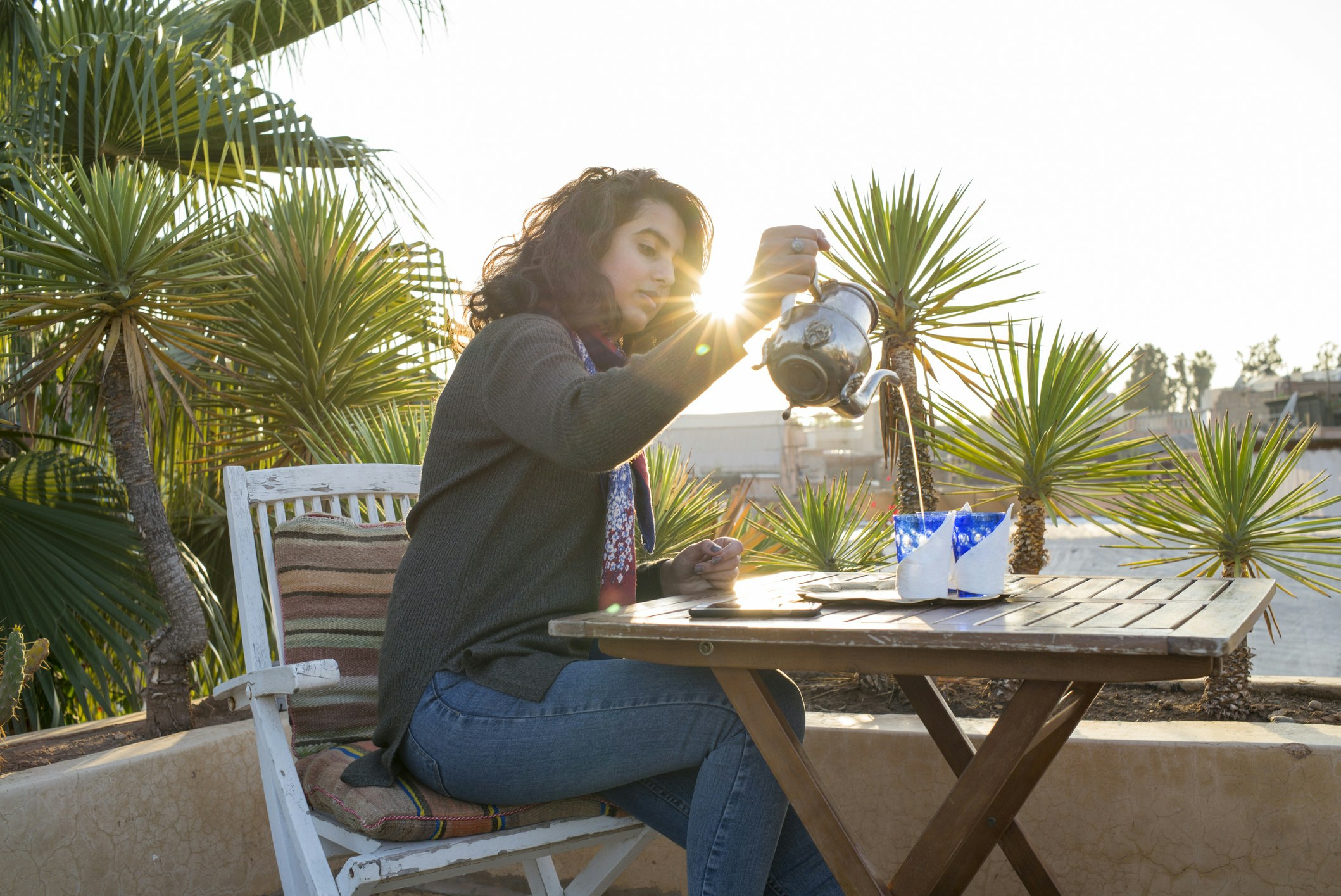 A woman with brown curly hair pours herself a cup of Moroccan mint tea in front of a low plaster wall and several short palm trees