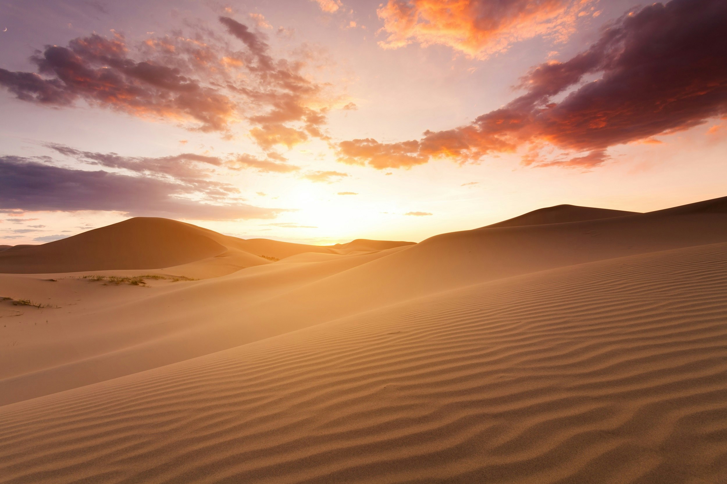 This image looks up the ripples of a large Saharna sand dune towards the setting sun; the sky is dotted with a few dark clouds, backed by a bright purple horizon.