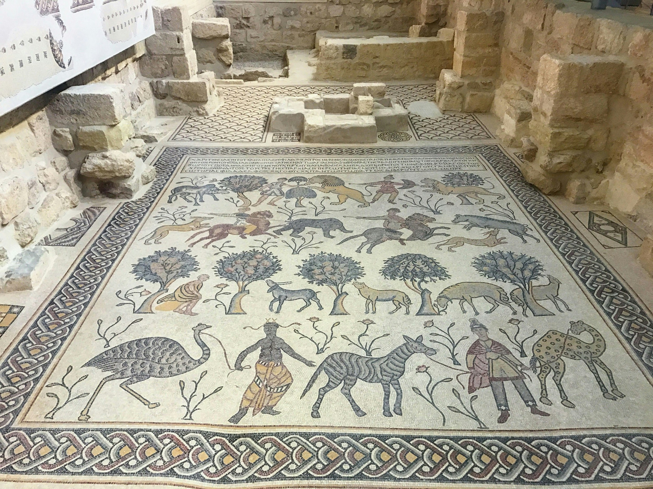 A lovely mosaic with braided border that features people, trees and various animals, such as zebras, ostriches, camels, donkeys and lions.