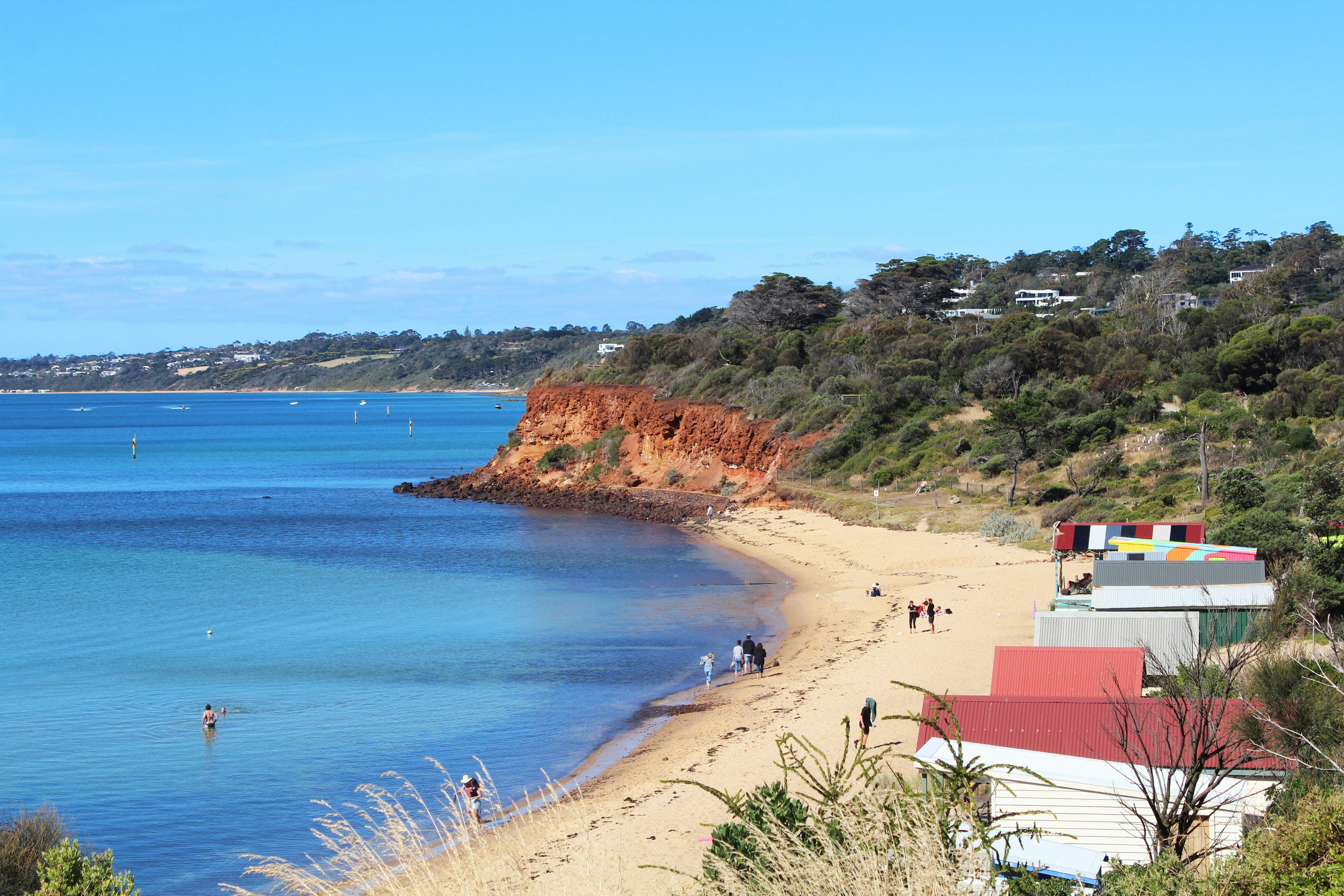 An elevated shot of Mornington’s Mothers Beach. A stretch of straw-coloured sand is sheltered in a small bay; several people are walking along the beach beside a few buildings, and a few people can be seen in the dark-blue water.