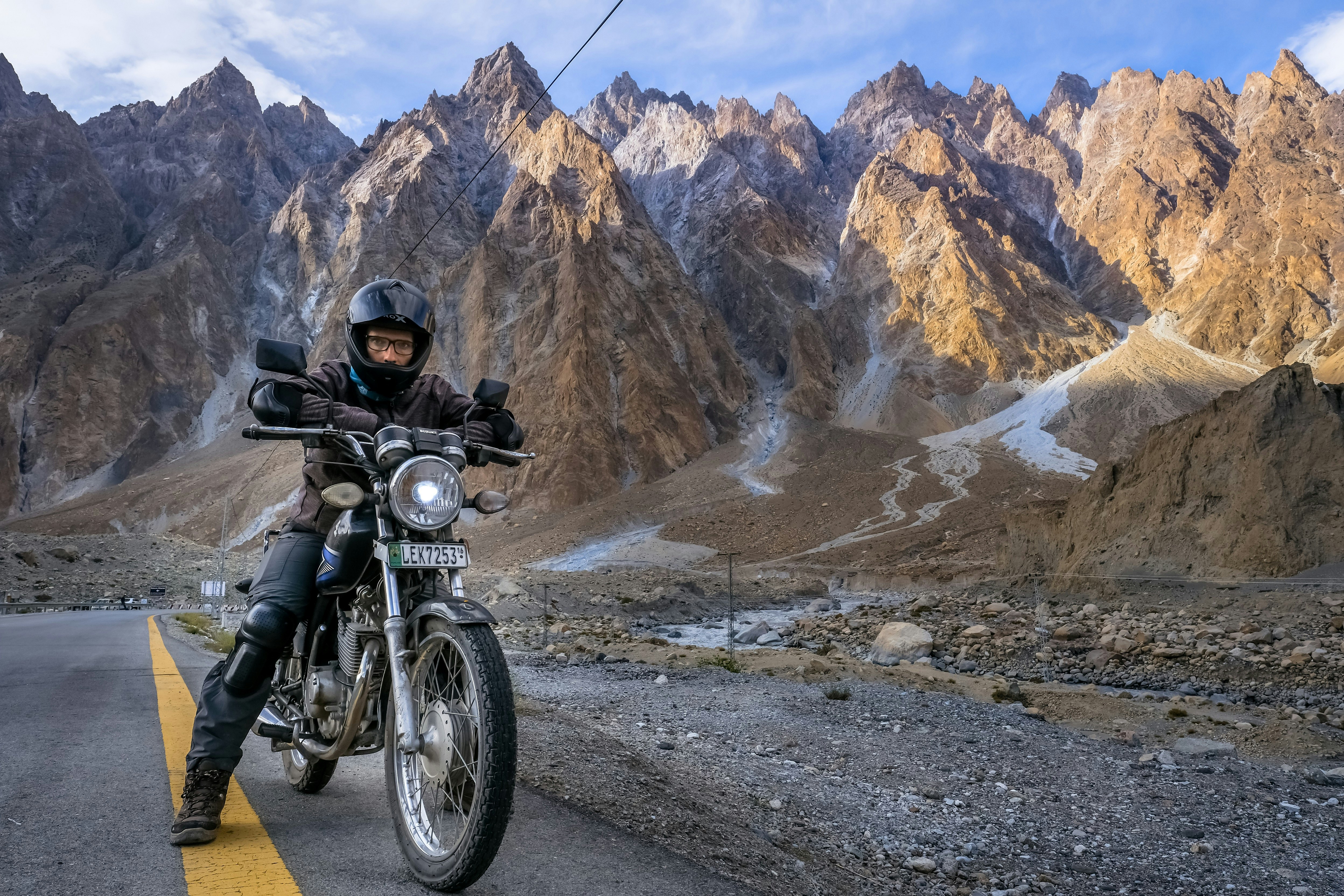 The author sits on his rented motorbike on the side of a road in Northern Pakistan. Behind him, a row of jagged mountain peaks are visible.
