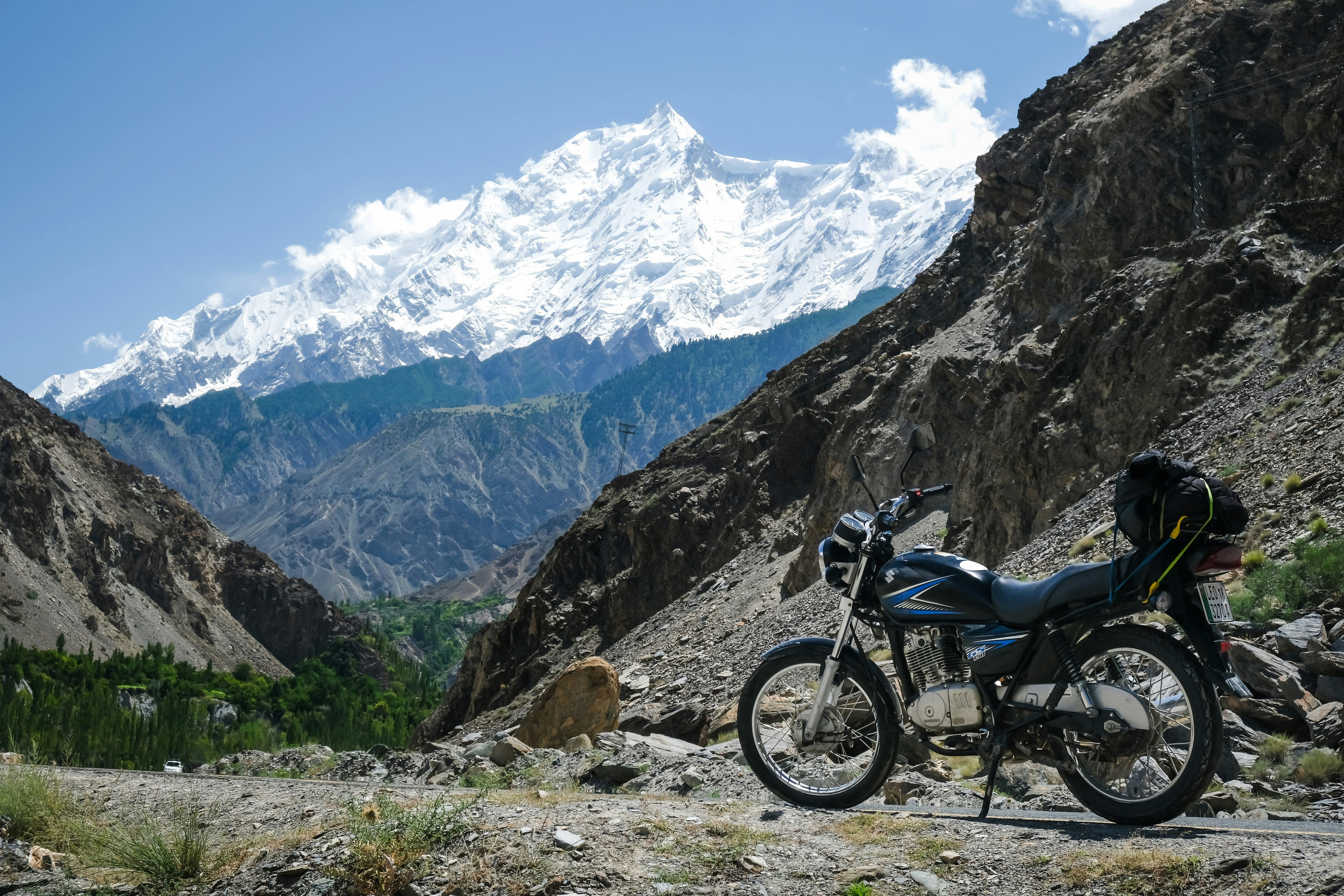 A motorbike stands next to a clearing in the mountains of Northern Pakistan. In the background, snow-covered mountains are visible.