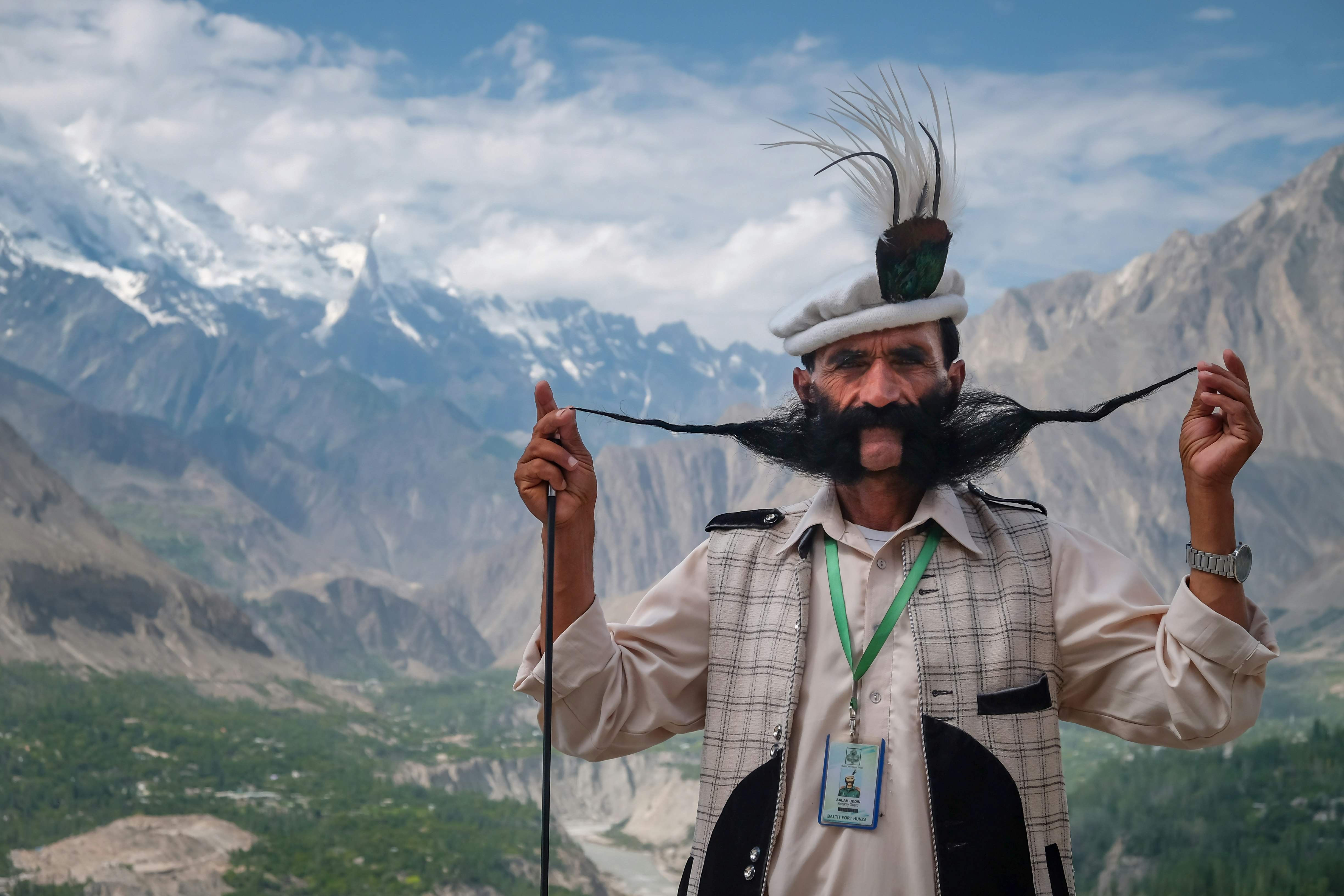 A local guide showcases his very long beard by holding the two ends up, away from his face. He is wearing a traditional hat, as well as a shirt and gilet. In the background a number of mountains can be seen.