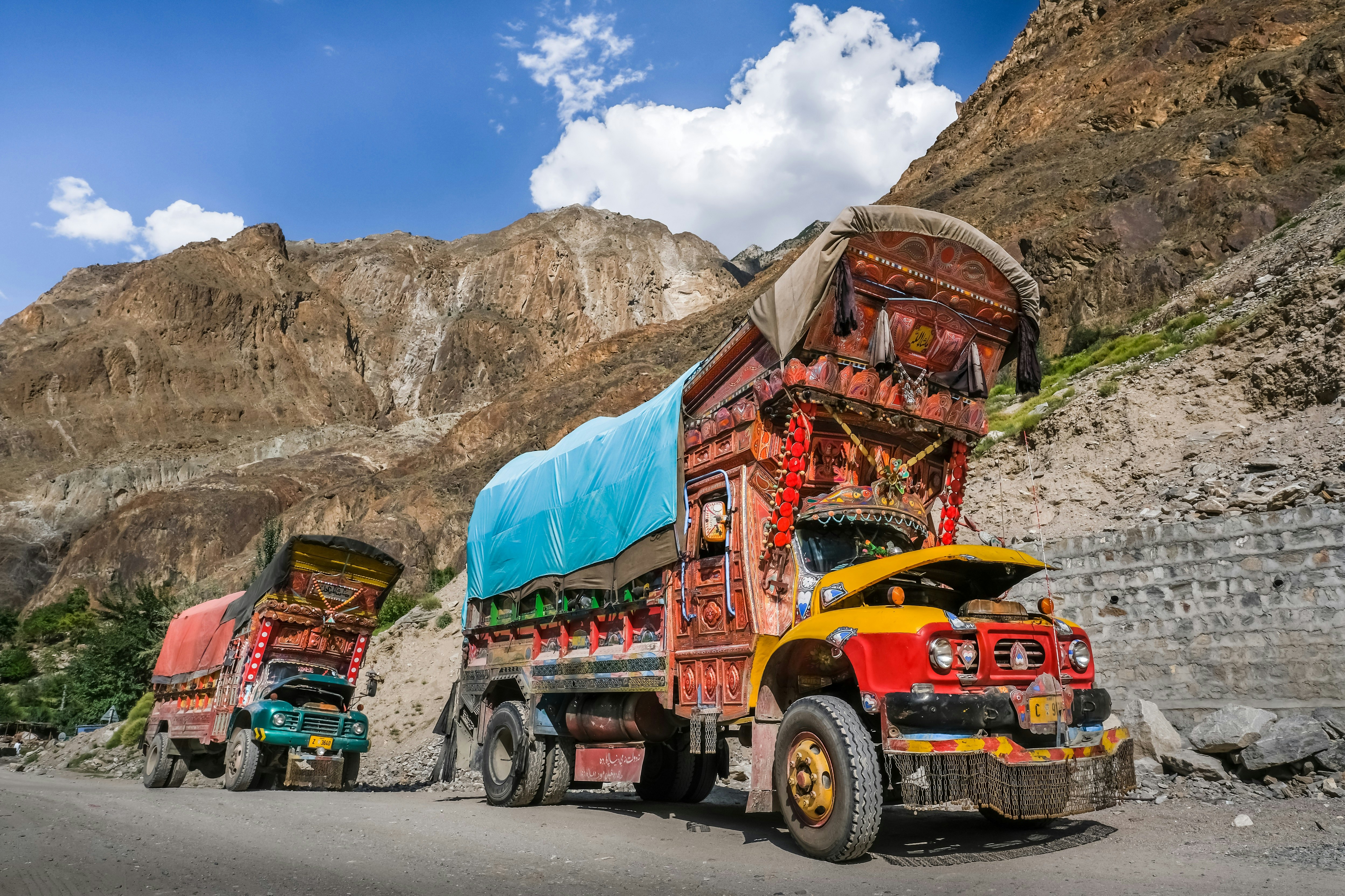 Two large lorries are parked on the side of a mountain road in Northern Pakistan. Both vehicles are lavishly decorated and adorned with flags, bells and tassels.