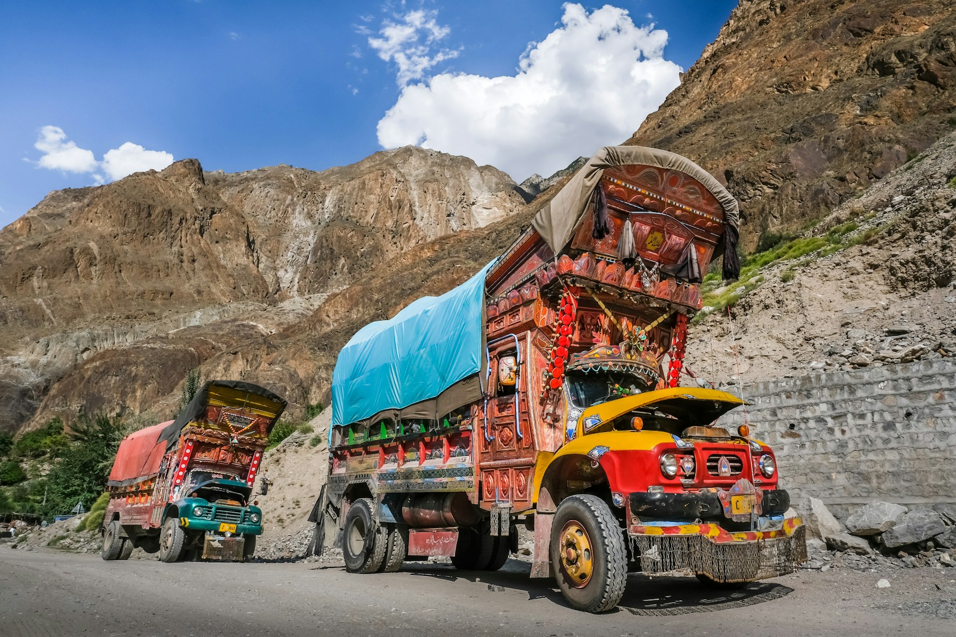 Two large lorries are parked on the side of a mountain road in Northern Pakistan. Both vehicles are lavishly decorated and adorned with flags, bells and tassels.