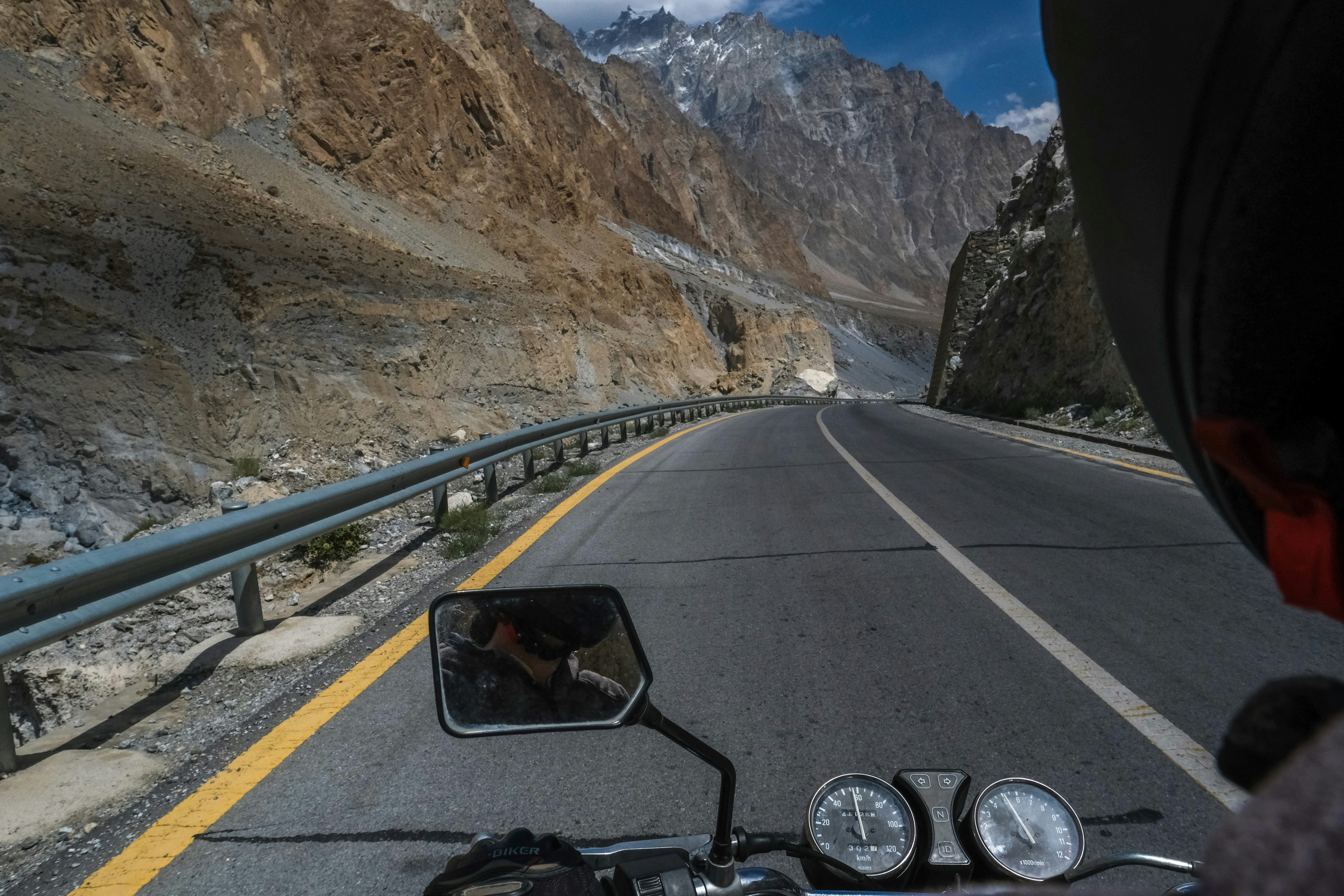 An over-the-shoulder shot of a motorcyclist driving on the roads of Northern Pakistan. The motorcycle's speedometer and wing mirror are visible in the foreground, while in the background, rocky mountains rise up on all sides.