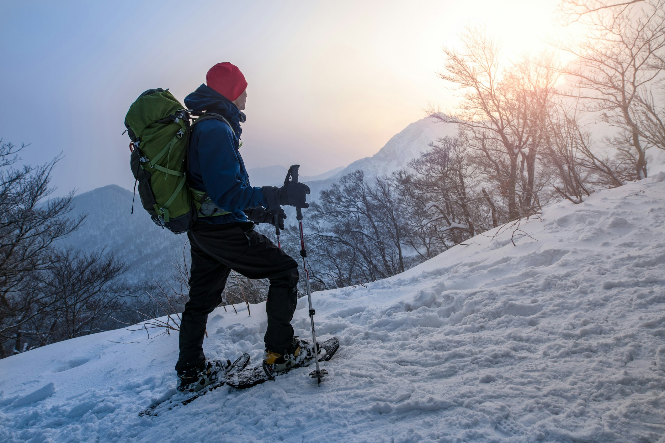 A man pauses as he snowshoes at sunrise on Mt. Daisen. He is wearing a red knit hat, a blue windproof ski jacket, has a olive green sixty liter backpack on, and is turned away form the viewer to admire the sun rising over a nearby peak