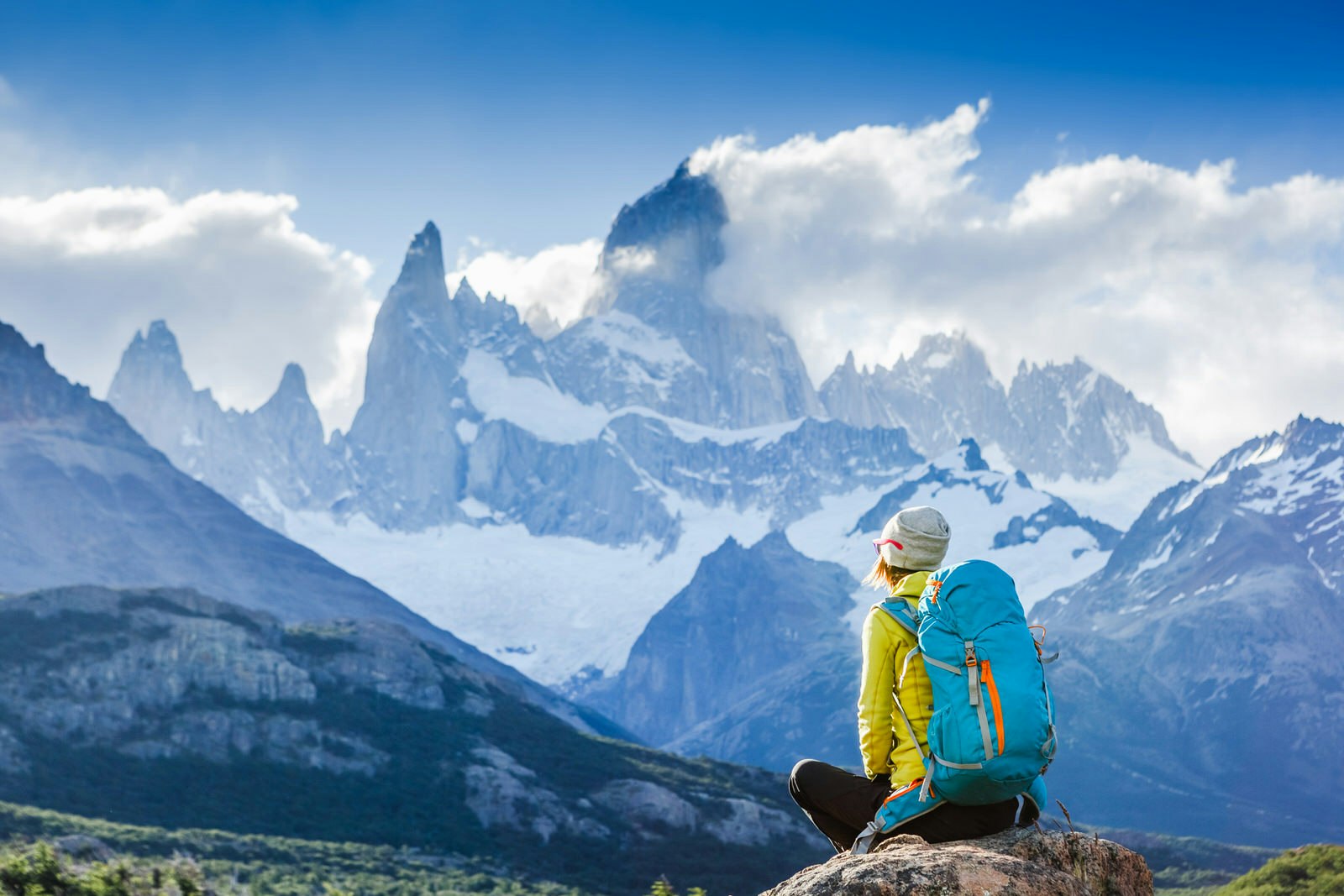 A woman in a yellow jacket, grey wool hat and blue backpack sits on a rock and looks up to the awe-inspiring jagged ridges and peaks of Mount Fitzroy.