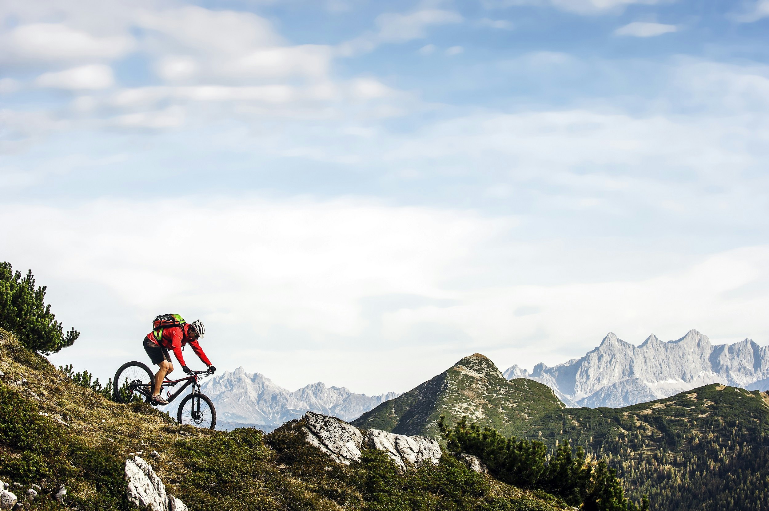A mountain biker descends a slope covered in grass, rocks and alpine vegetation; in the background is a green hill, which is backed in the distance by a jagged ridge of rocky Alps peaks.