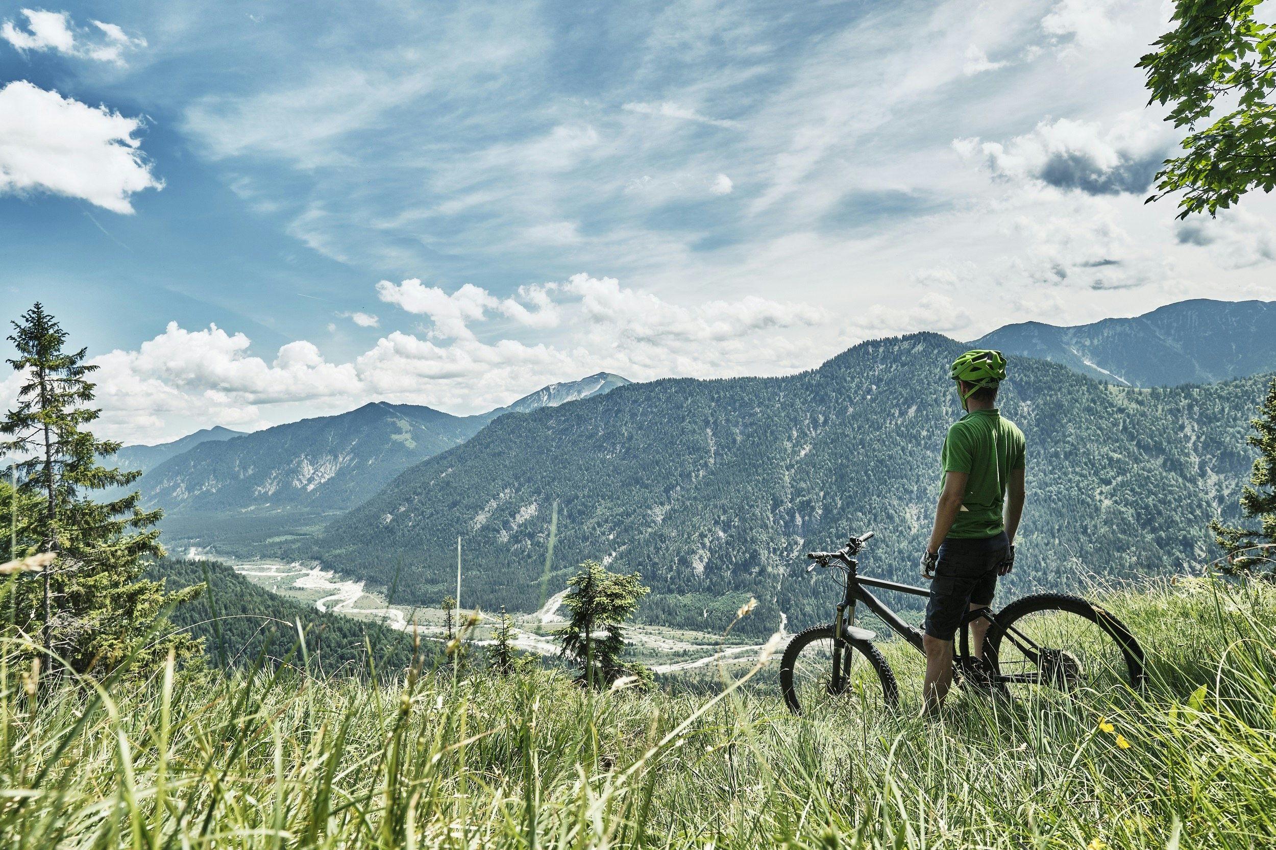 A mountain biker stands over his bike on a long grassy slope and looks down over the river flowing in the distant valley floor.