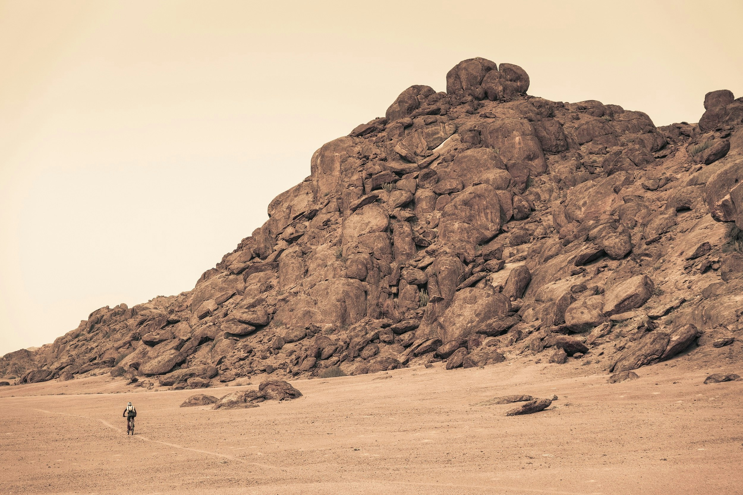 A lone mountain biker rides across a desert landscape past a large rock outcrop that towers above; everything is hued in brown tones. 	