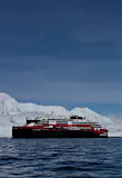 The Hurtigruten hybrid cruise ship MS Roald Amundsen sits on calm waters beside vast icebergs in Antarctica on a clear day.
