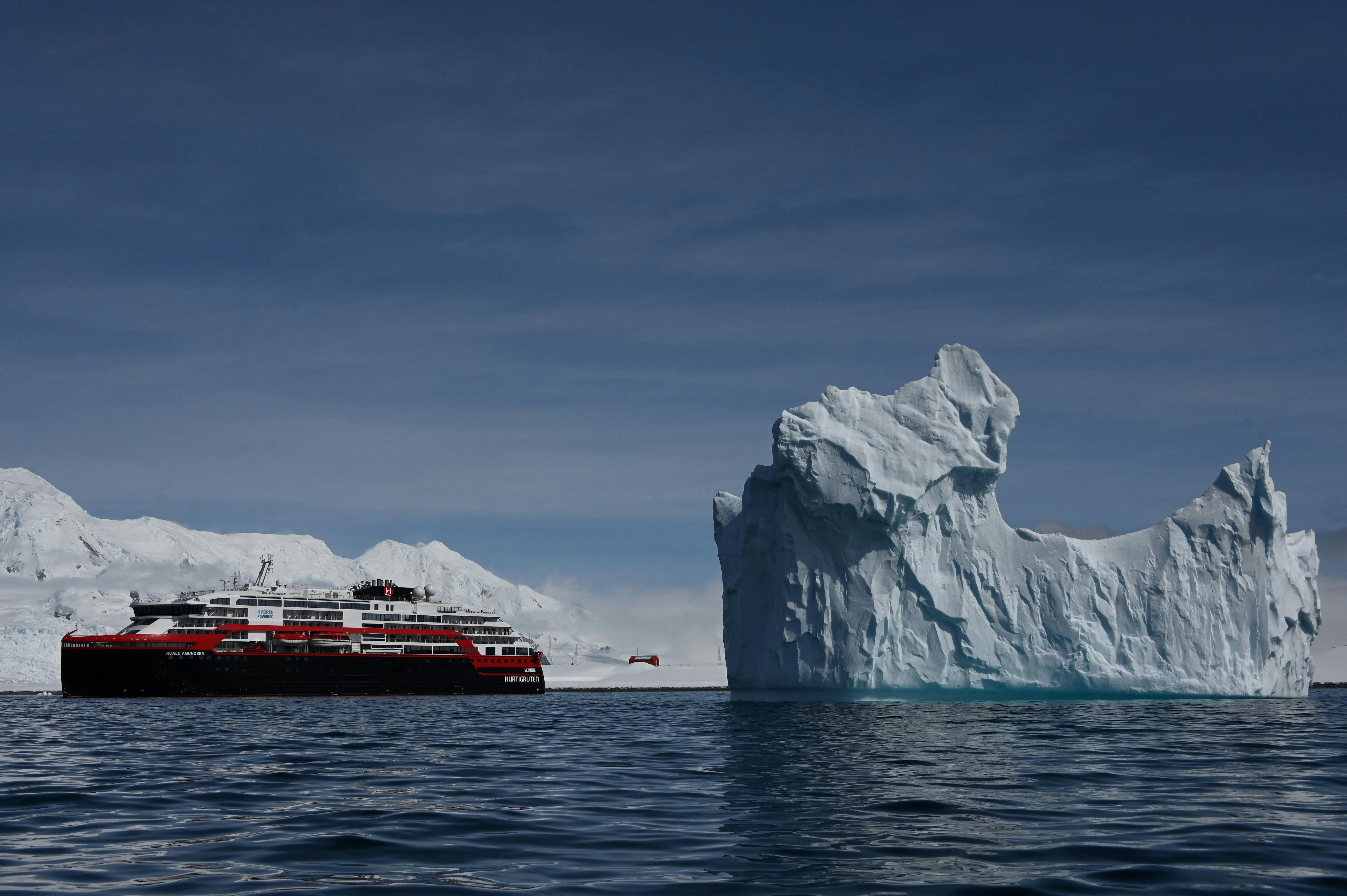 The Hurtigruten hybrid cruise ship MS Roald Amundsen sits on calm waters beside vast icebergs in Antarctica on a clear day.