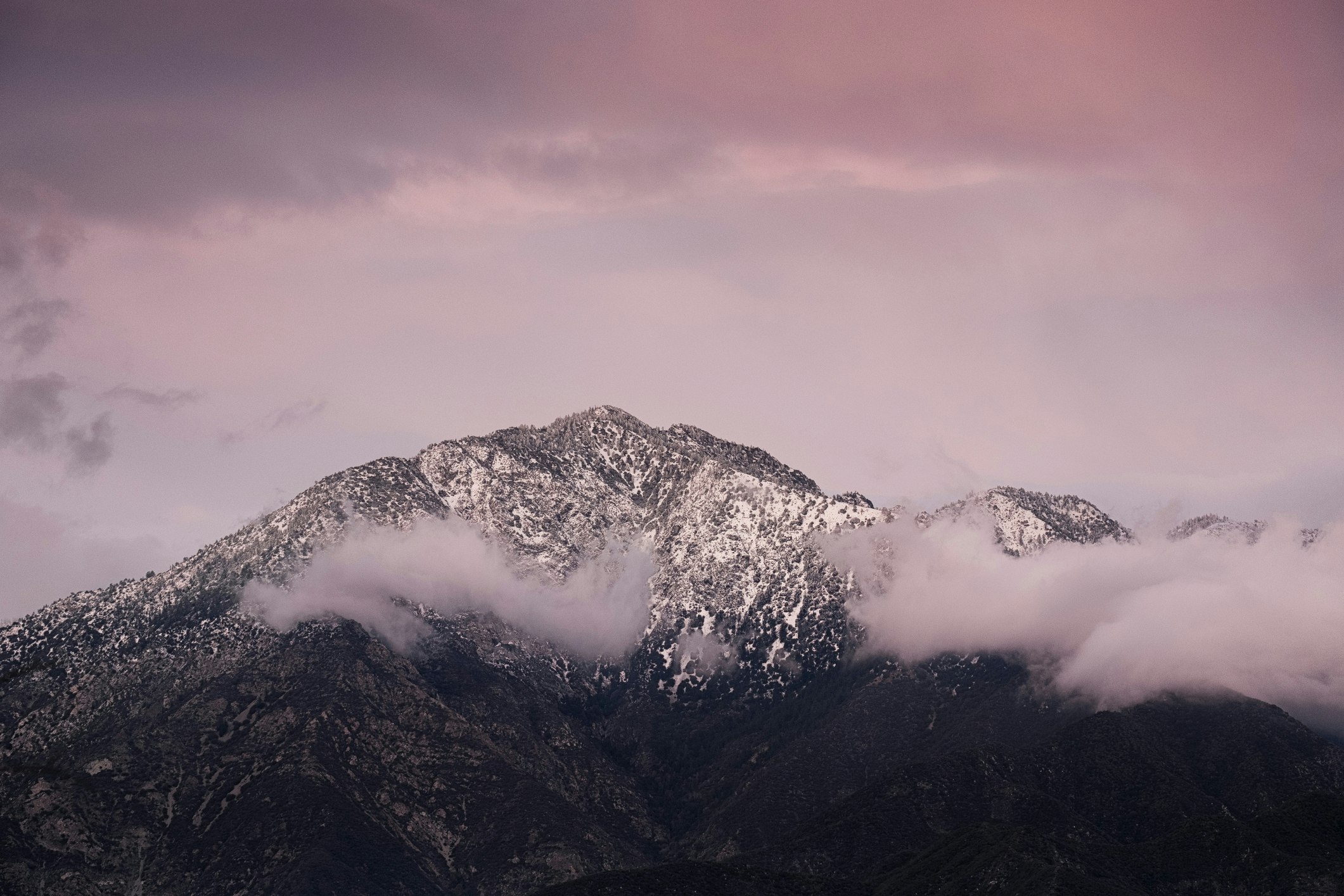 The grey, snow-dusted summit of Mt. Waterman in Angeles National Forest in the San Gabriel Mountains overlooking Los Angeles is ringed with wispy clouds tinted pink by the sunset, which lights the whole sky in pastel lavenders and dusty rose, and contrasts with the dark shadow of the mountain in the lower third of the shot, 