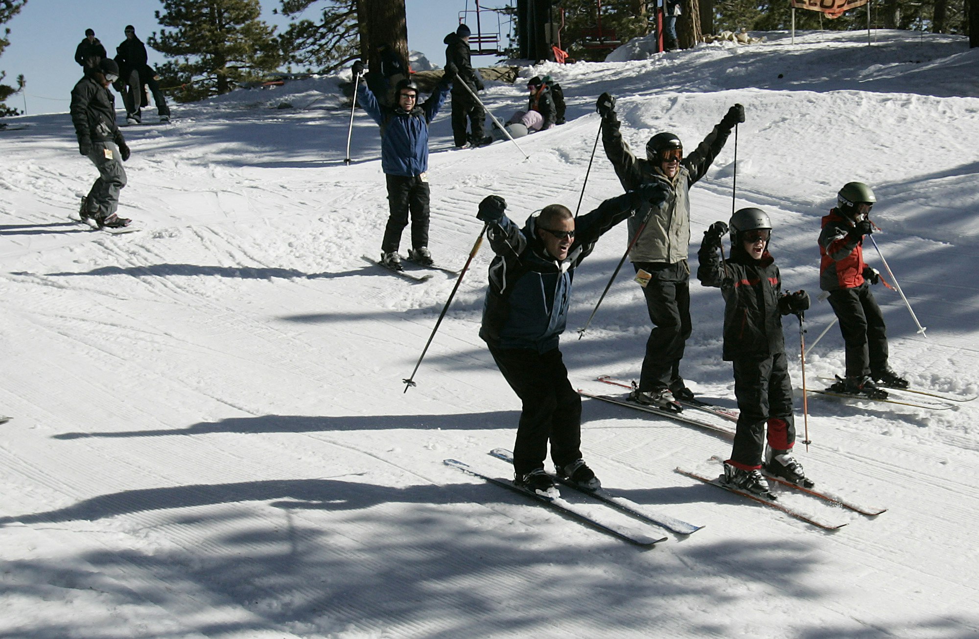 Two men and two boys in dark colored ski apparel raise their hands and ski poles in excitement as they coast down the slope of Mt. Waterman on a groomed ski trail