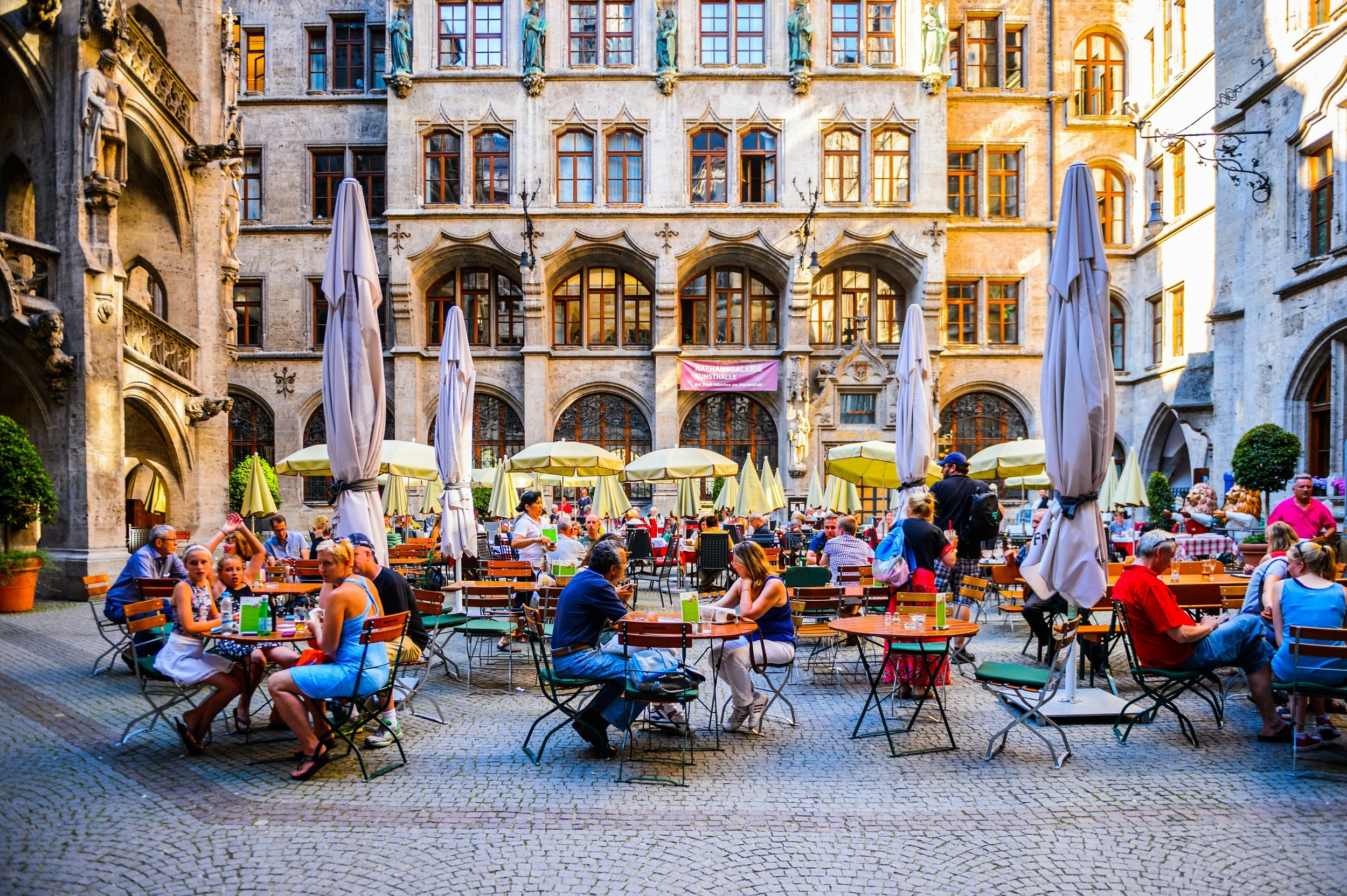An open-air cobbled square, surrounded on three sides by tall stone buildings, is full of people sat at tables with umbrellas.