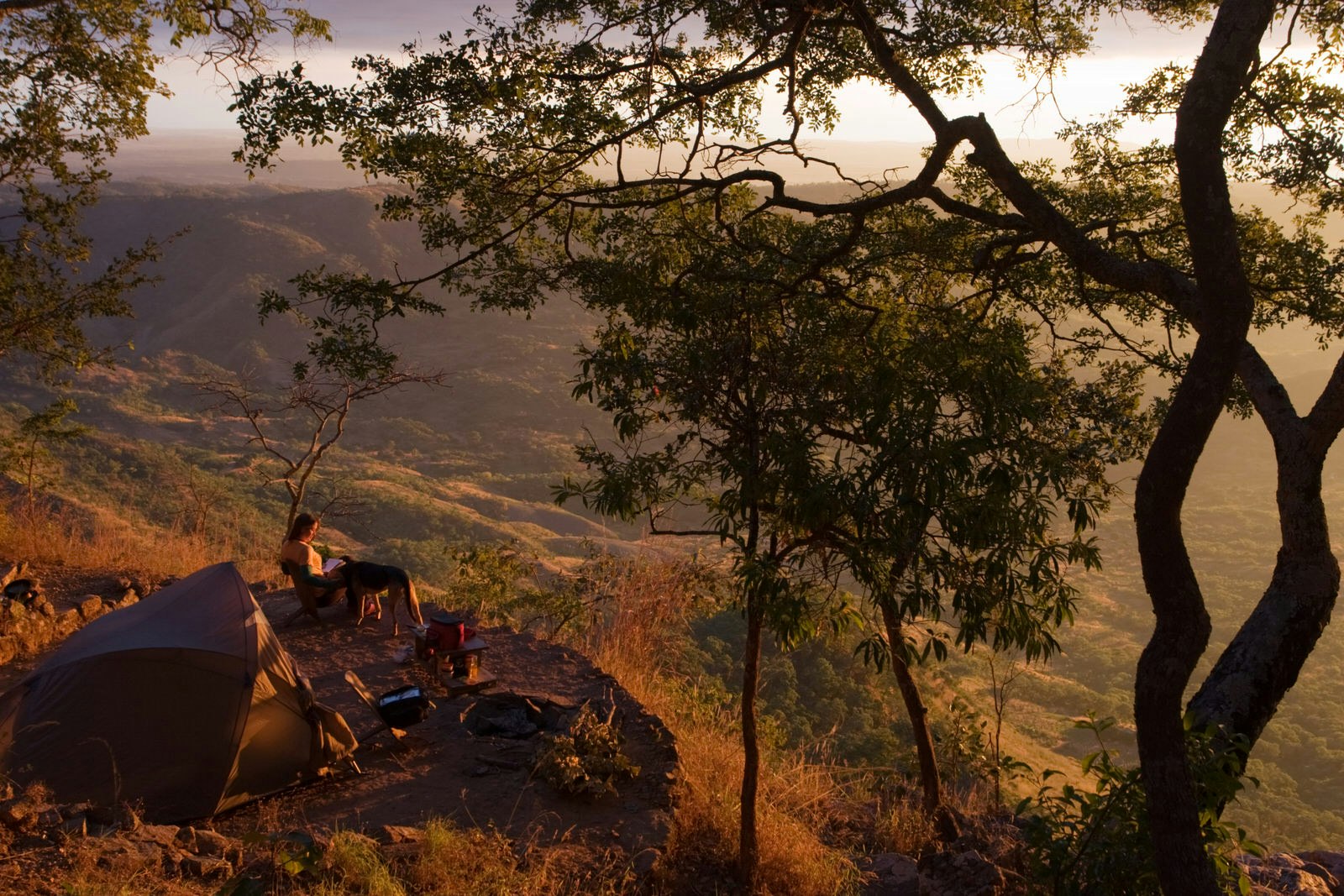 A woman sits in a camp chair next to her dog on a flat, circular campsite plot this is perched on the side of a steep slope overlooking the valley far below; the light is golden, likely at sunset