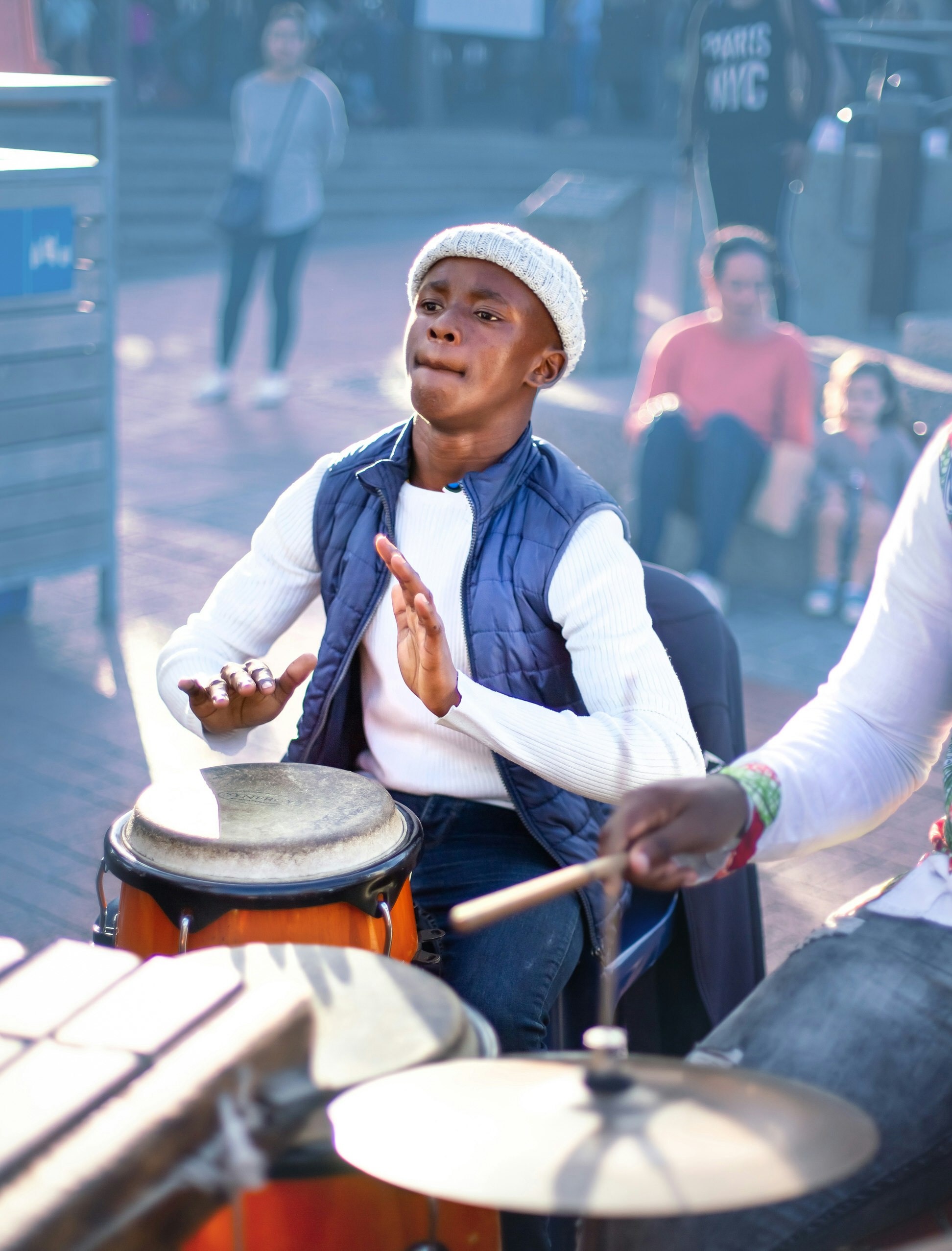 A young musician plays a traditional African drum, with a blurred set of modern drums in the foreground.