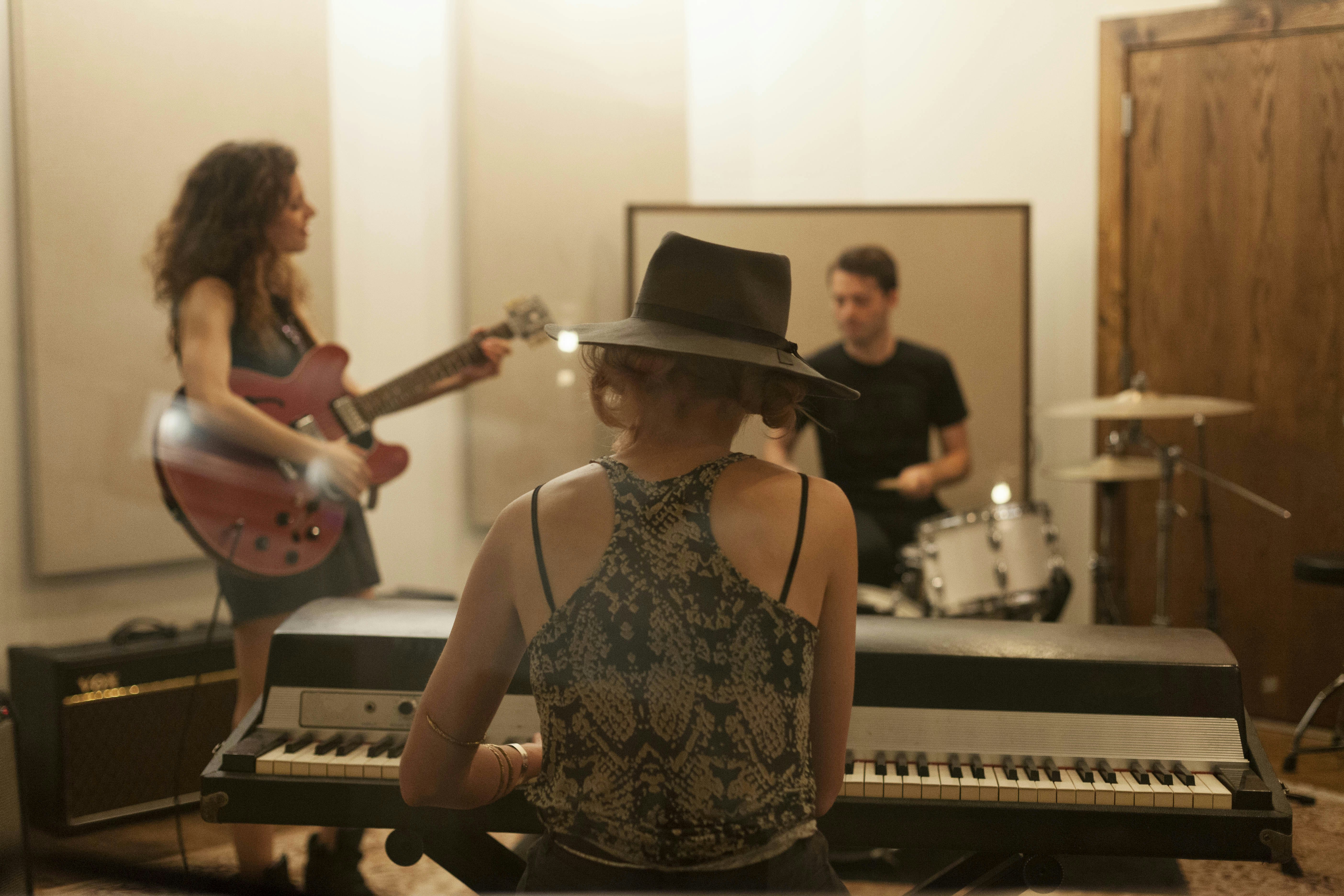 Musicians playing. A woman playing the piano has her back to the camera. To her left a woman is playing the guitar and in front of her is a man playing the drums 