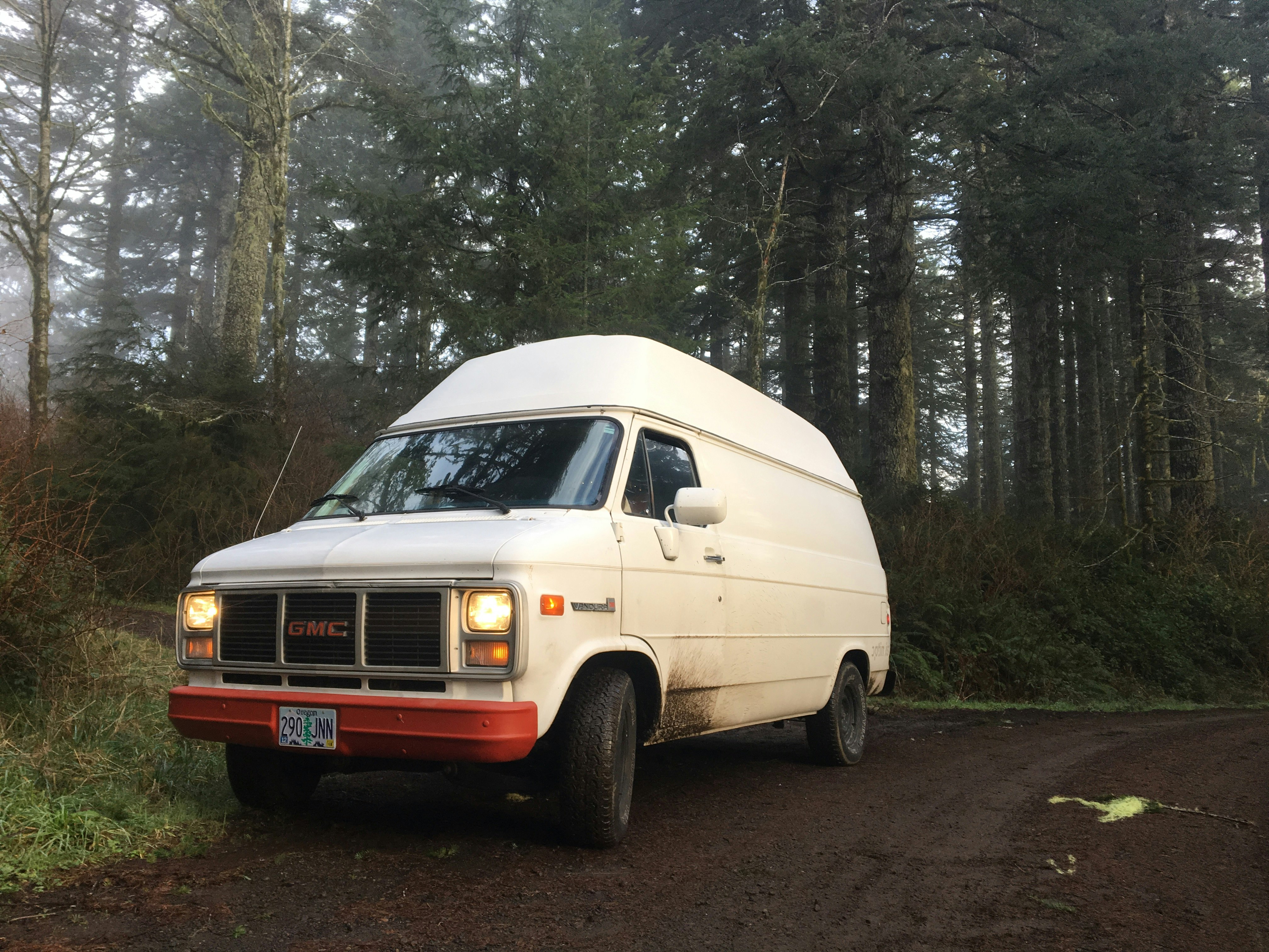 A white 1986 GMC Vandura van with a cherry red bumper sits on the edge of a dark brown dirt road in a forest in Oregon. Spruce and pine trees stand in the background with fog rolling in on the left side of the photo.