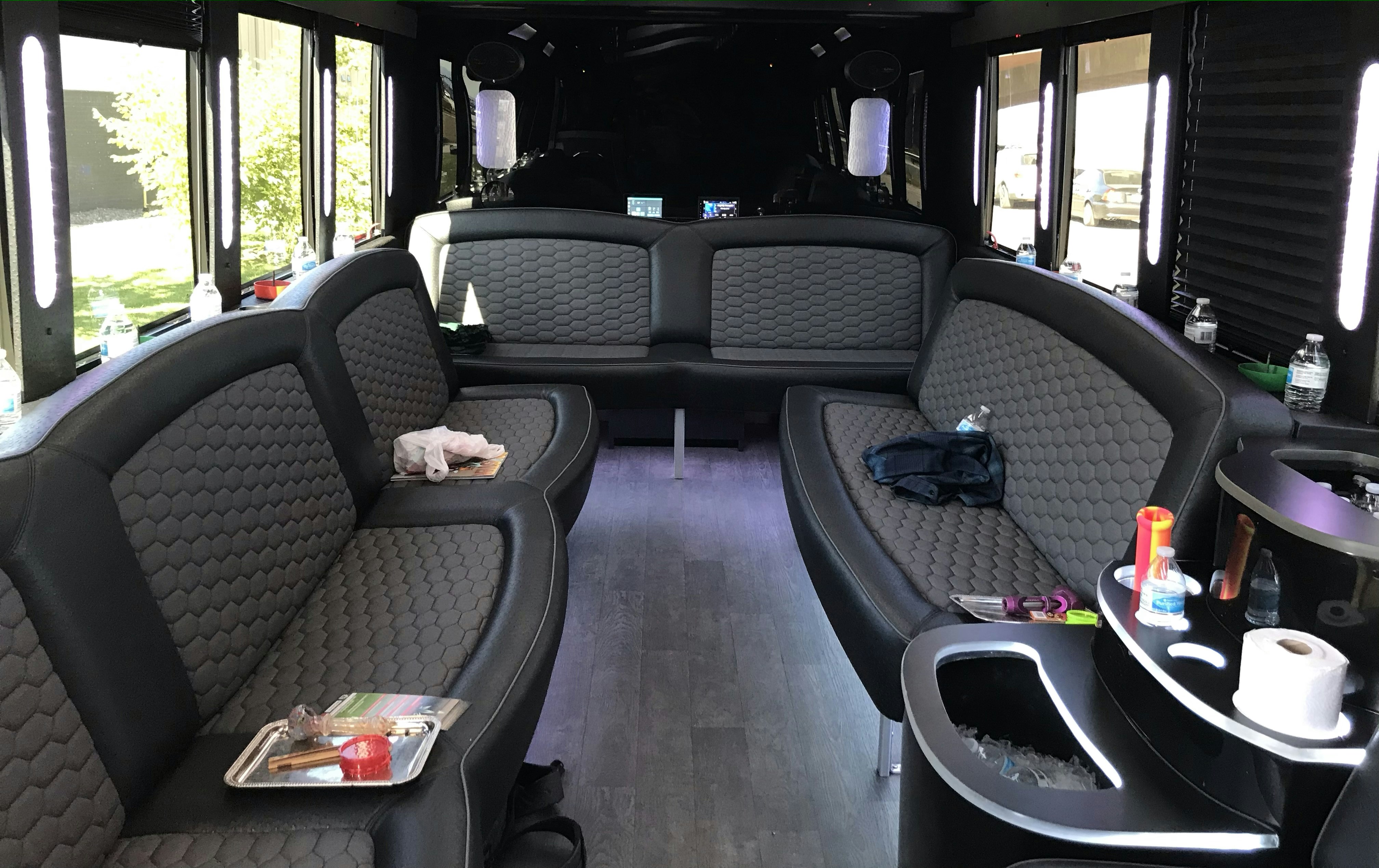The black and grey interior of the My 420 Tour bus, where the upholstered seats have small metal trays laid out with cannabis paraphernalia and bottles of water and tucked on various surfaces. 