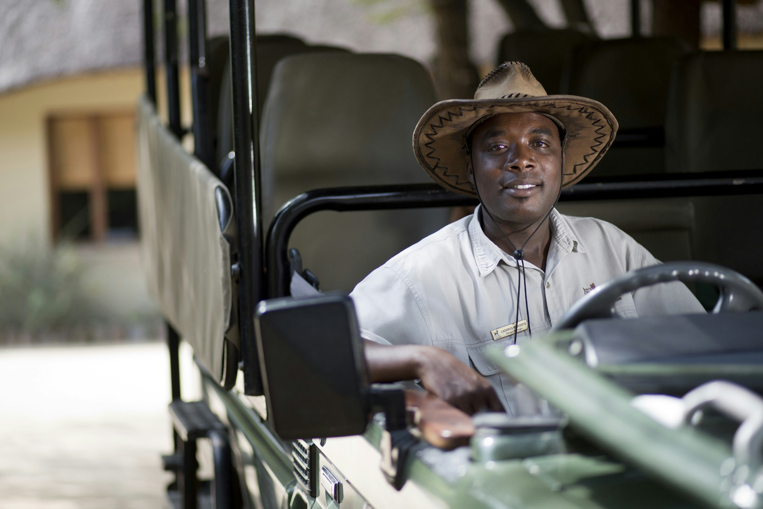 A park ranger safari guide sits in the driver seat of an empty 4WD wildlife drive vehicle; behind him are three rows of empty seats beneath a canvas canopy (there are no doors or sides to the vehicle).