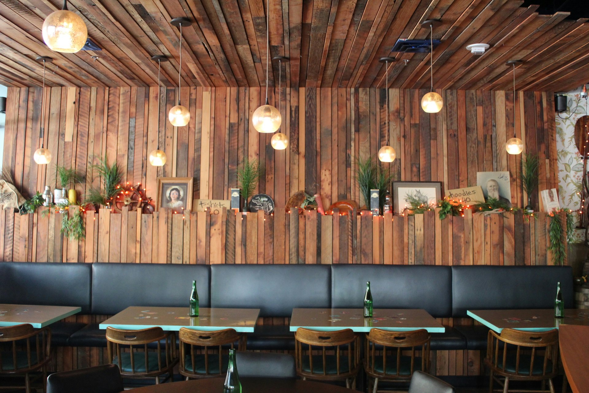 The interior of Navy Strength, a tiki bar in Seattle, is lined with worn wood panels from the top of the black banquet seating to the ceiling, which is also lined. White round lamps hang from the ceiling, and a shelf displays framed art, tchotkes, sea grass, and other ephemera   