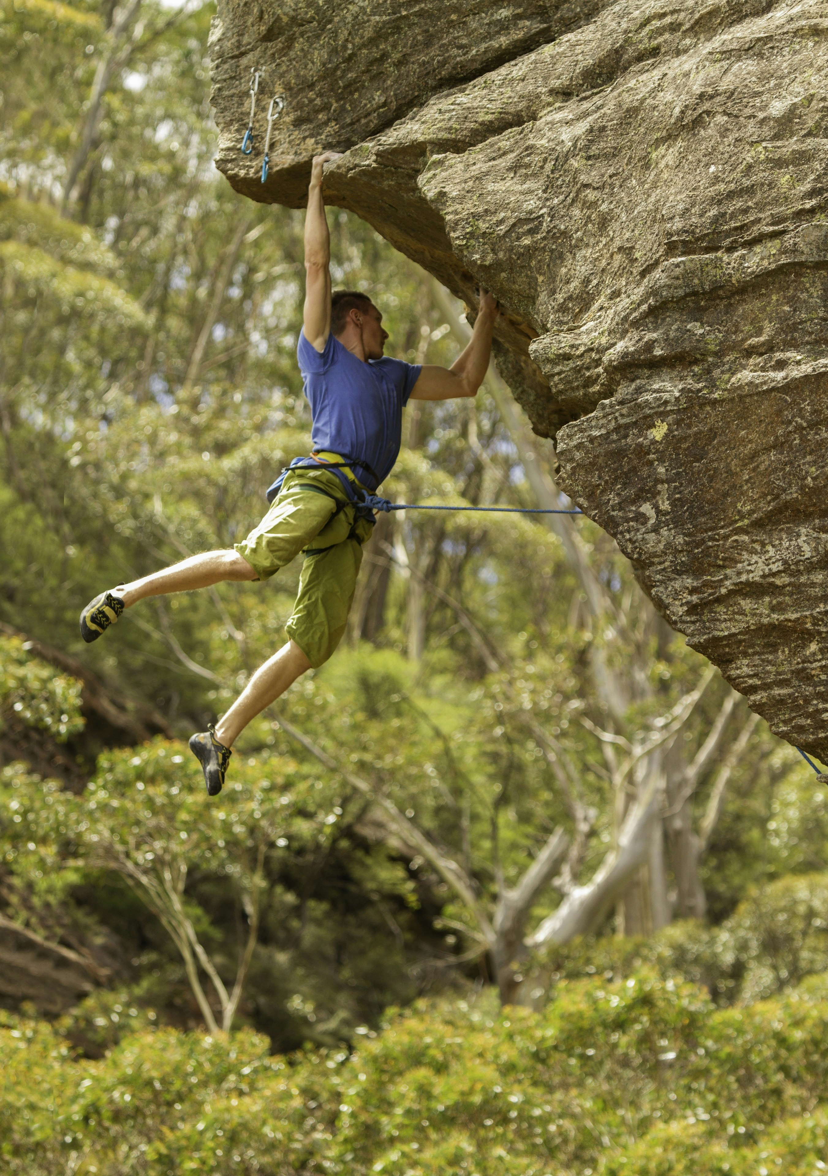 A rock climber hangs from his hands below a rock overhang; a rope from his harness leads to the rock. Behind is a lush forest setting.