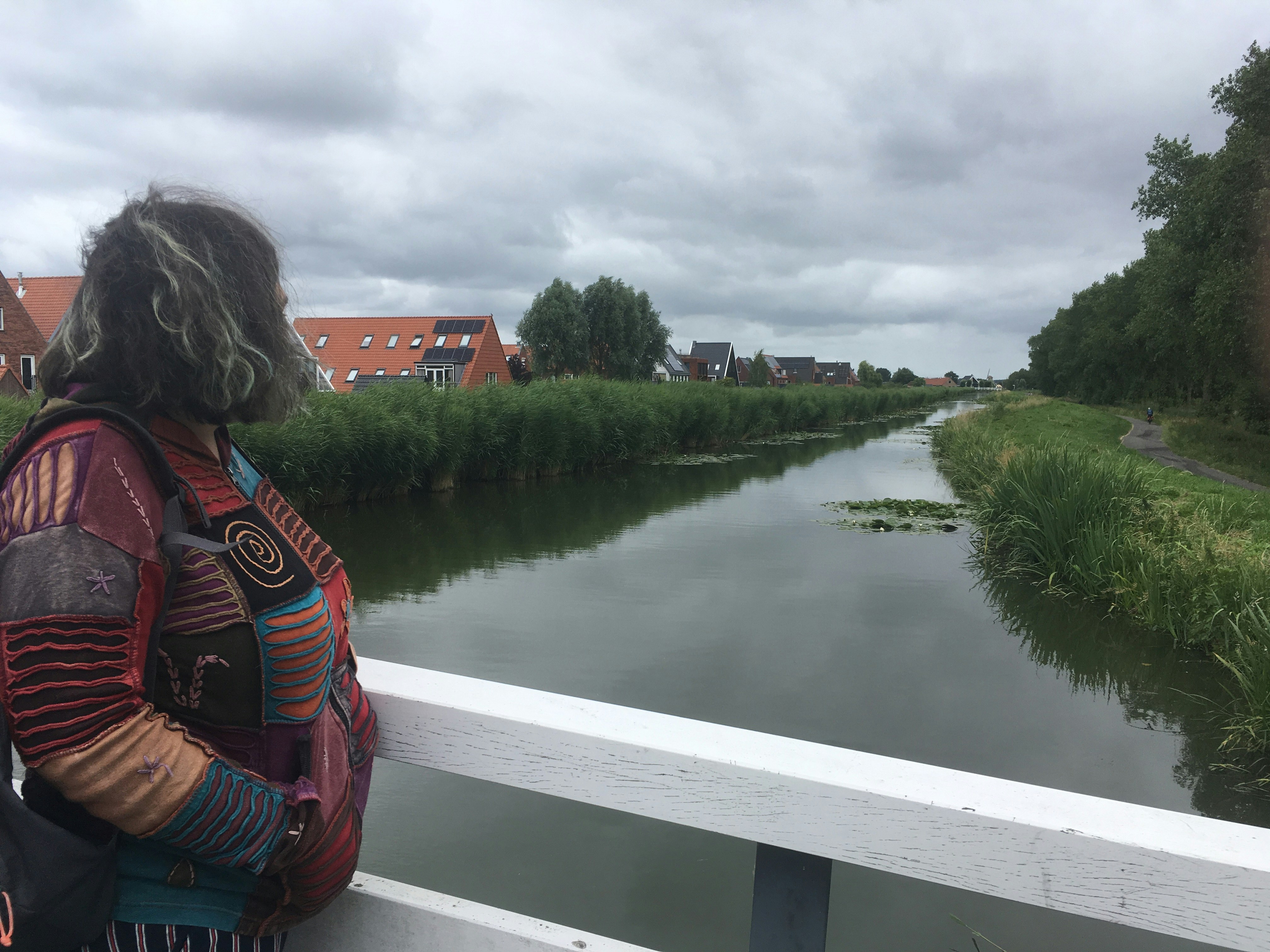 A woman stands on a bridge turned away from the camera. She's looking at the view of a long river with houses on one side and a path on the other