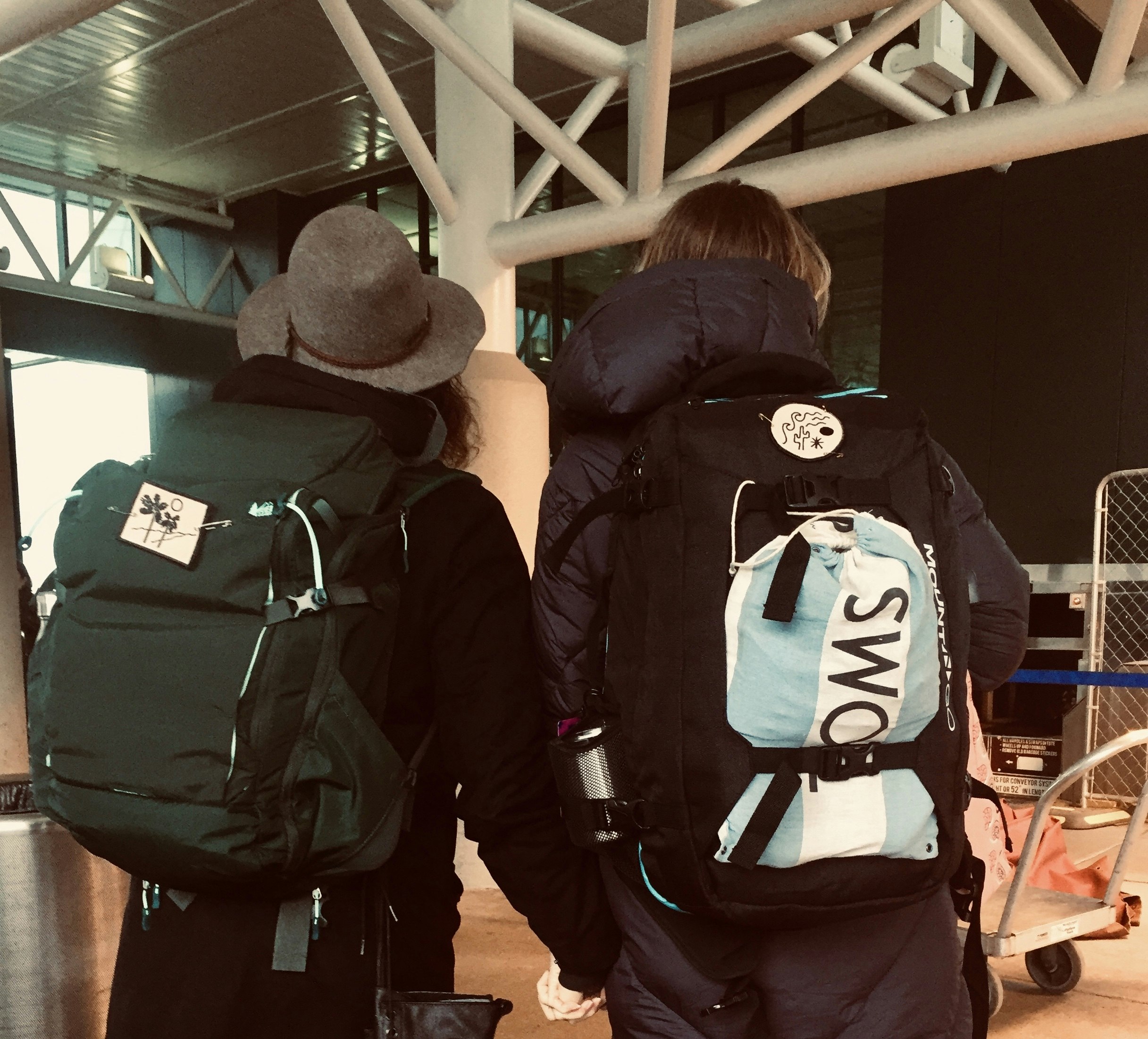 Nik and Lindsey stand with their backs to the camera, both wearing coats and carrying backpacks. They are holding hands, and are facing the entrance to the Newark Airport