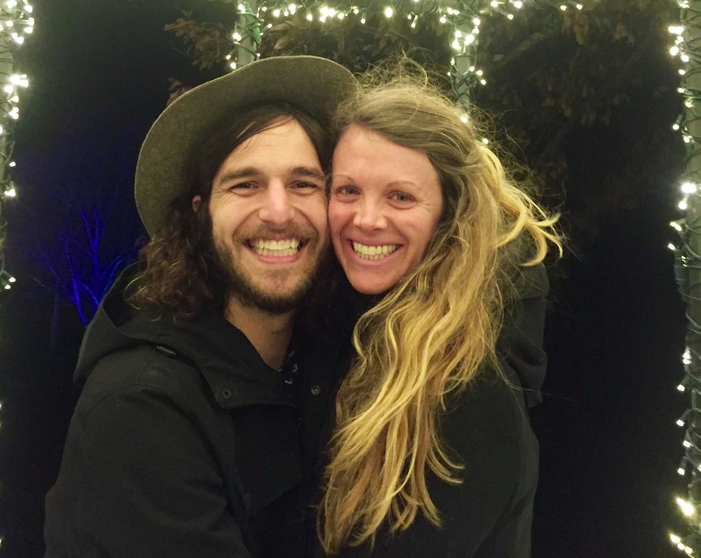 A couple with huge, genuine smiles stand under a gazebo coated in white Christmas lights. Nik is on the viewer's left in a grey hat and black jacket with long curly brown hair to his shoulders and a short beard. Lindsey has very long blond wavy hair to the bottom of the frame. She is wearing a black jacket.