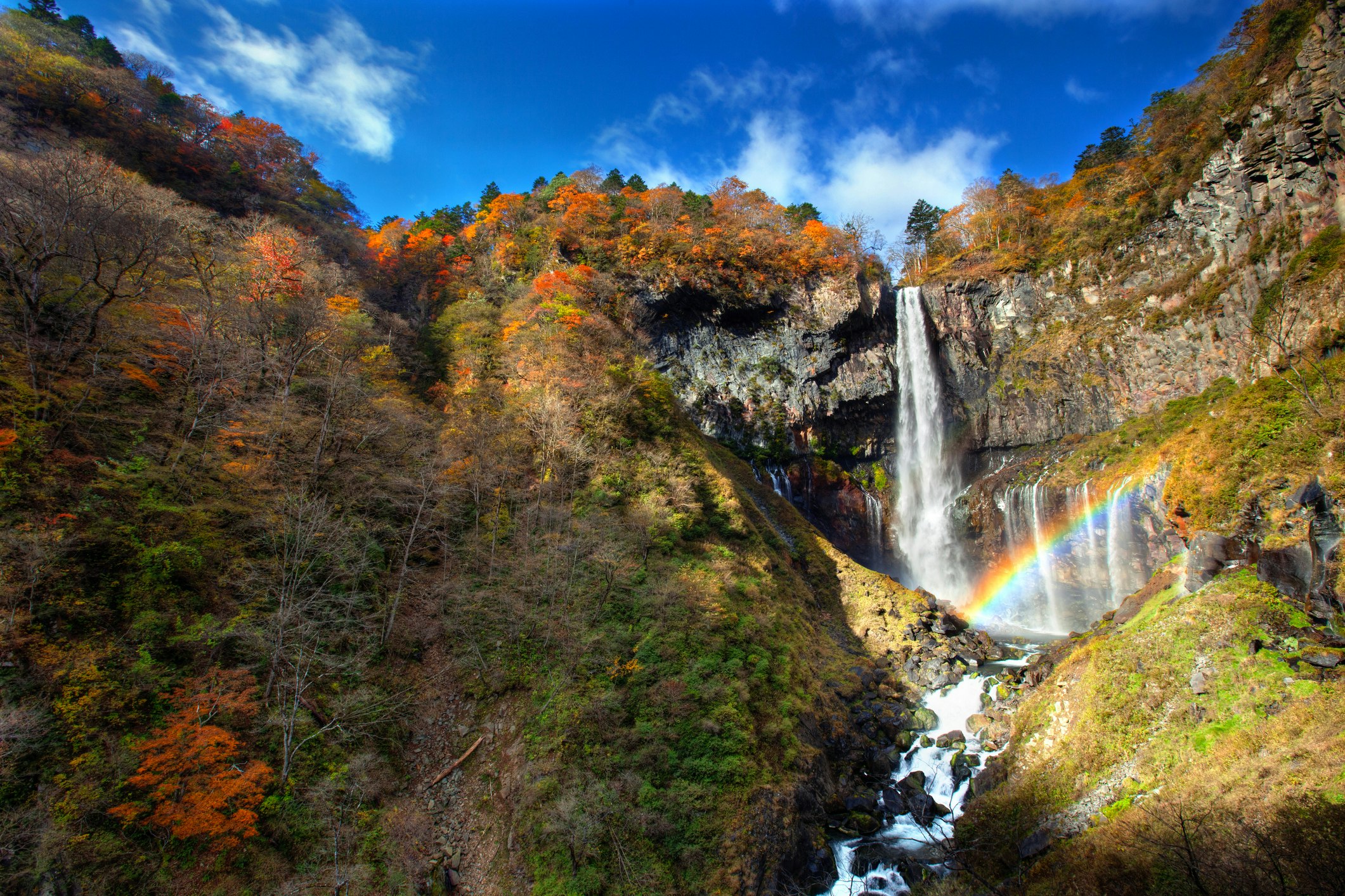 Kegon Falls spills through a crack in the volcanic mountain face of Nikko National Park, which are covered in autumn foliage. A rainbow is formed in the mid ground 