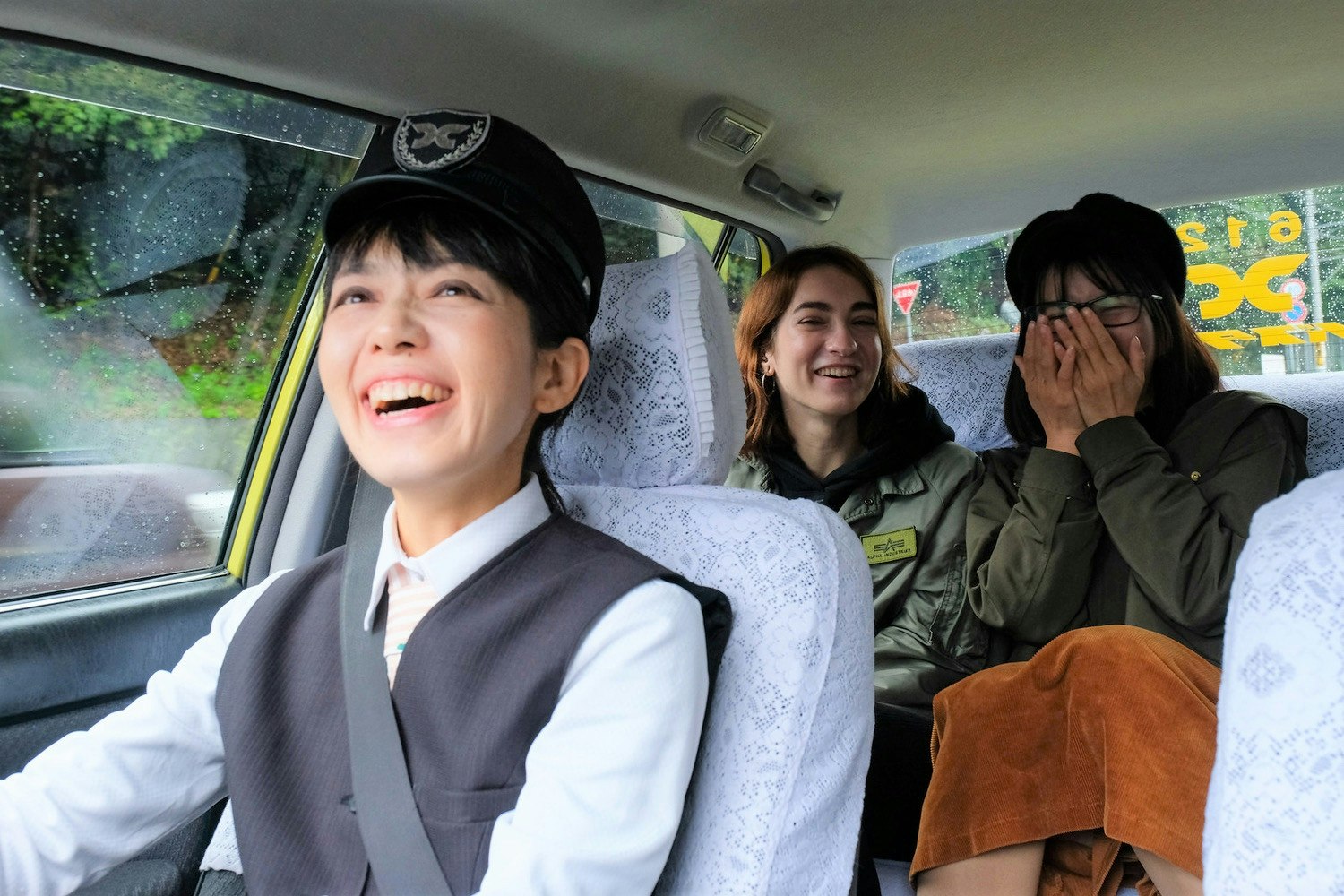 A laughing udon taxi driver in a conductor's hat and a grey vest looks in the rear view mirror at her giggling passengers in the back seat, who are wearing olive green jackets and an orange skirt. The taxi's seats are covered in lace doily slip covers and a light rain is falling on the windows outside
