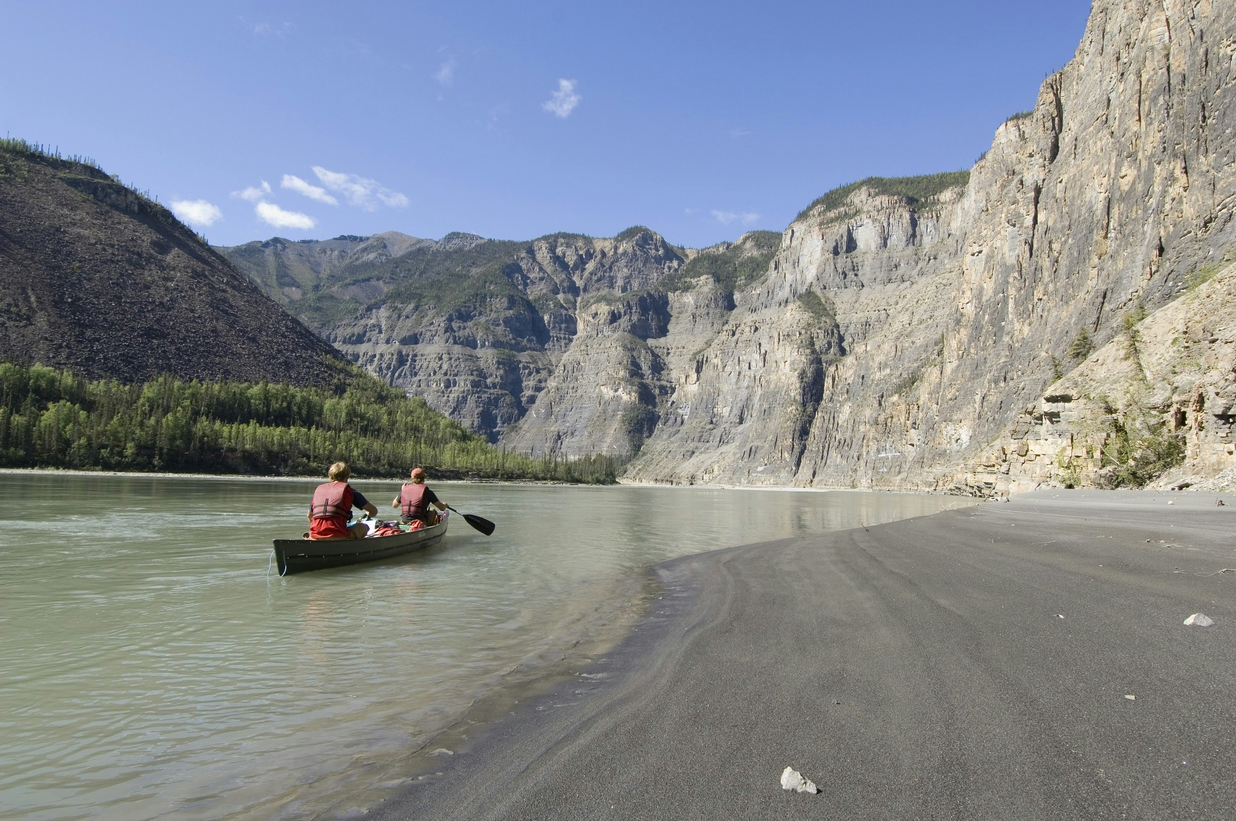 People in a canoe paddling through a canyon of the South Nahanni River, Northwest-Territories; they are close to a grey beach (to their right), with forested shoreline on the left; ahead are steep rocky bluffs that cascade down into the river.