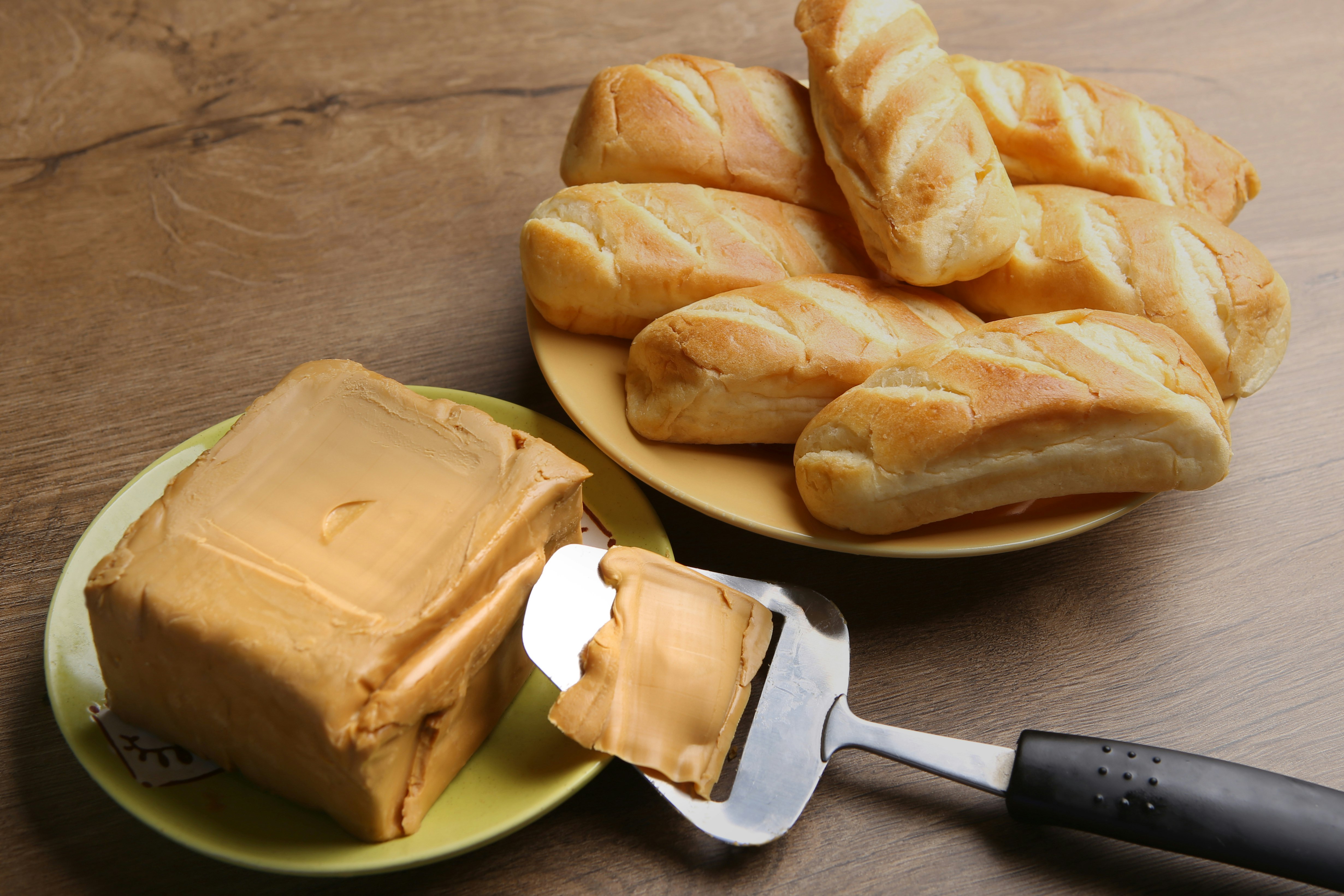 A large, light brown brick of brunost sits on a pistachio green plate with a cheese slicer resting against it with a thin strip of brunost freshly cut. In the background is a plate of hard Norewegian dinner rolls, all on a warm wood table.