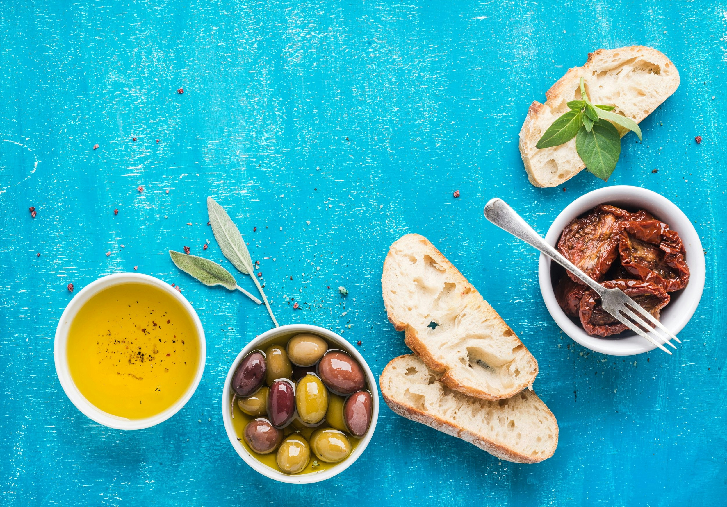 On a bright azure-coloured table sits a number of bowls; one holds olive oil, another olives and the final one sun-dried tomatoes; some slices of bread are also on the table.