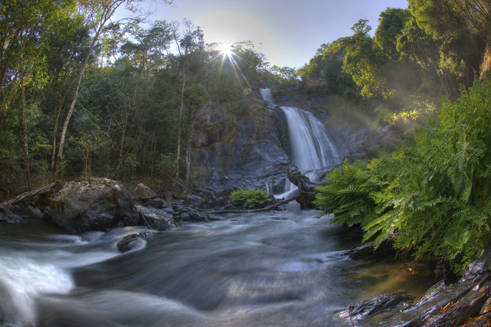 A shot looking up along a fast flowing river to a huge, two-stepped waterfall; surrounding it all is lush forest, with the sun poking through the trees above the cascading water