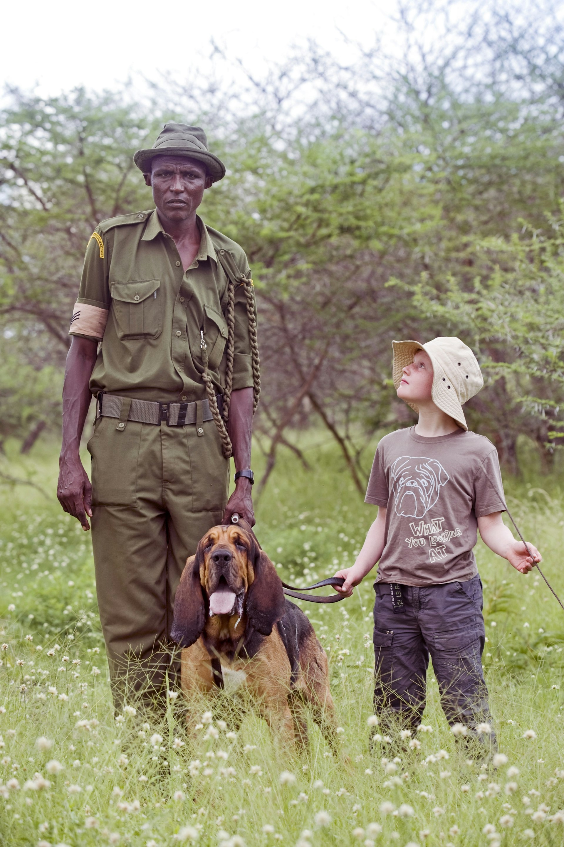 An anti-poaching ranger stands in uniform holding the lead of his bloodhound; a young tourist boy stands next to the dog and looks up to the ranger.