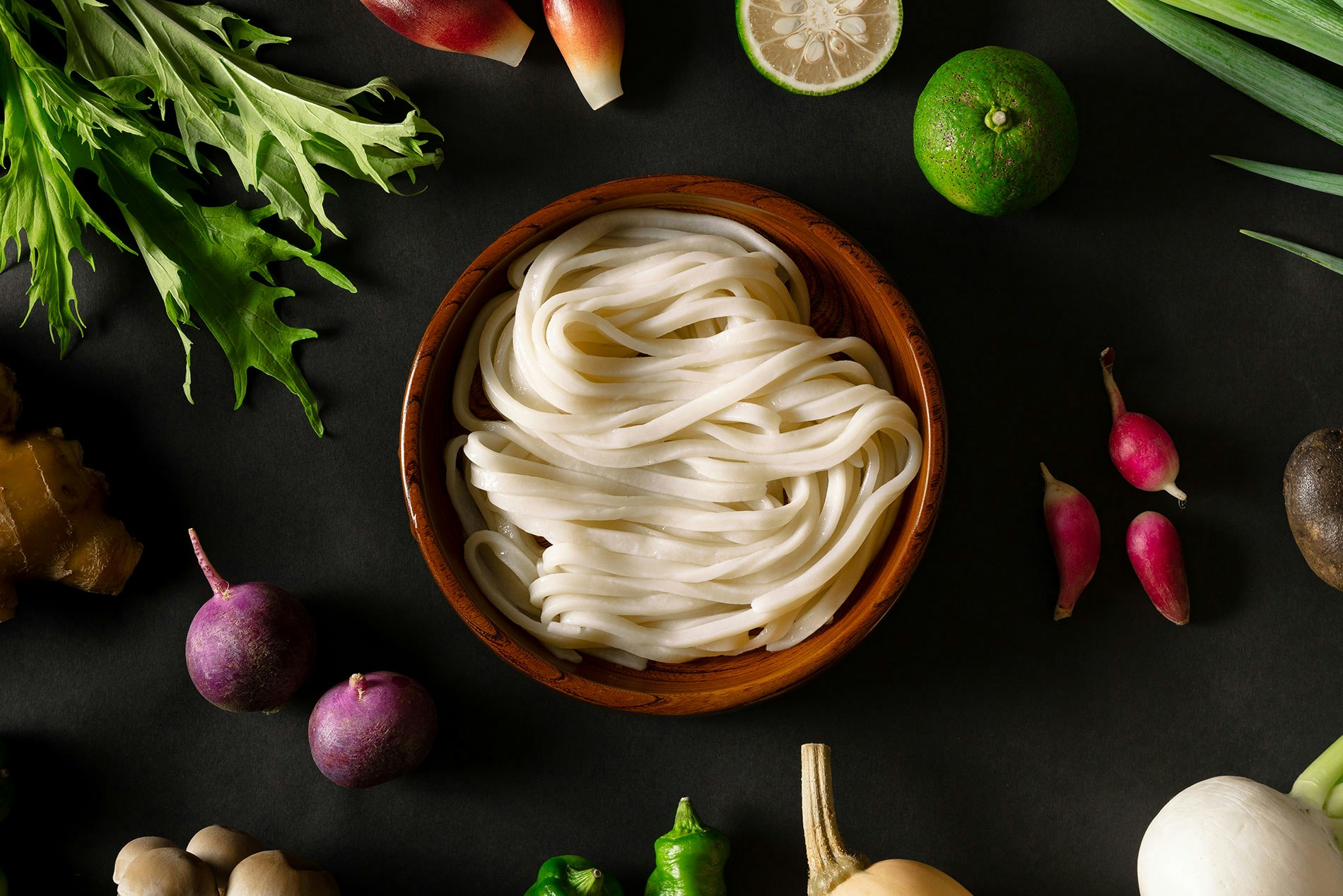 A bowl of noodles sits on a black table; it's surrounded by well-placed fresh vegetables.