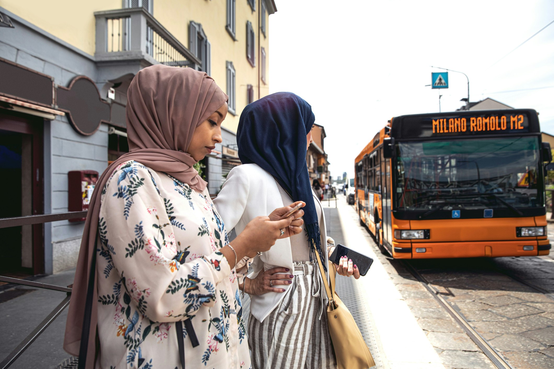 A pair of Muslim women, one in a mauve head scarf and the other in navy blue, turn slightly away from the camera as they wait for a bus in Milano, Italy which is pulling up on the right side of the frame